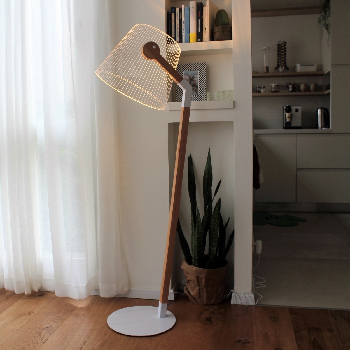 Ein stilvolles Zimmer mit einer modernen Flurlampe Set ZIGGI-B ausgestattet. Die Lampe hat einen Holzstamm und eine weiße Basis. Sie steht auf einem Holzboden neben einem Schreibtisch, Bücherregal, Zimmerpflanzen und hellen Vorhängen, die natürliches Licht einlassen.