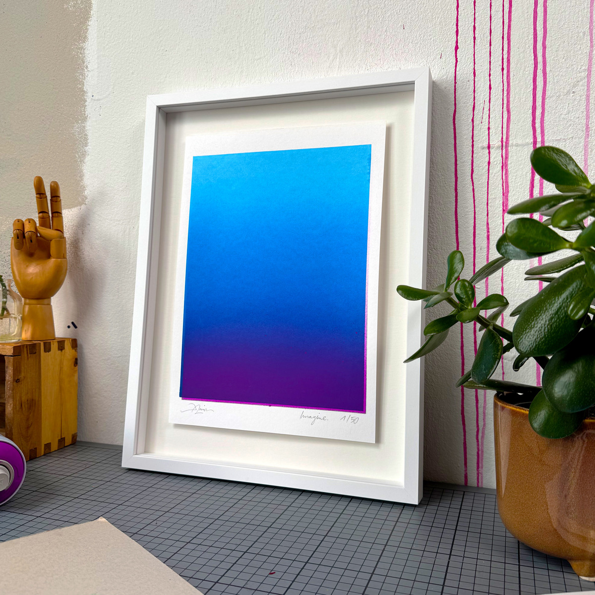 Gerahmtes Kunstwerk von Daniel Lisson, Titel »Imagine« (Blue/Violet), im Format 40x30 cm. Das Motiv zeigt einen sanften Farbverlauf von kräftigem Blau zu Violett, gerahmt in weißem Eichenholz, angelehnt an eine Wand. Im Vordergrund stehen eine hölzerne Hand, eine Pflanze und weitere Deko-Elemente.