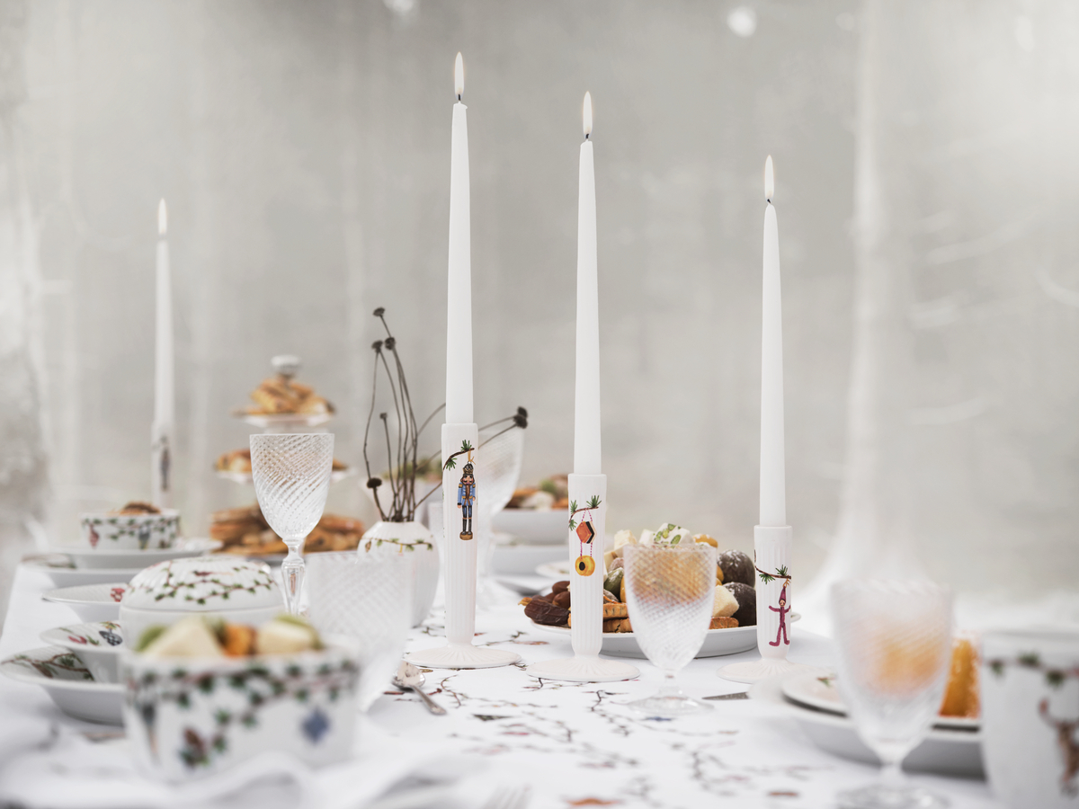 Ein festlich gedeckter Tisch mit mehreren schlanken weißen Kerzen in eleganten »Hammershøi«-Kerzenständern. Die Kerzenständer sind schlicht und modern gestaltet. Im Hintergrund sind unscharf dekorative Elemente und Geschirr zu sehen.