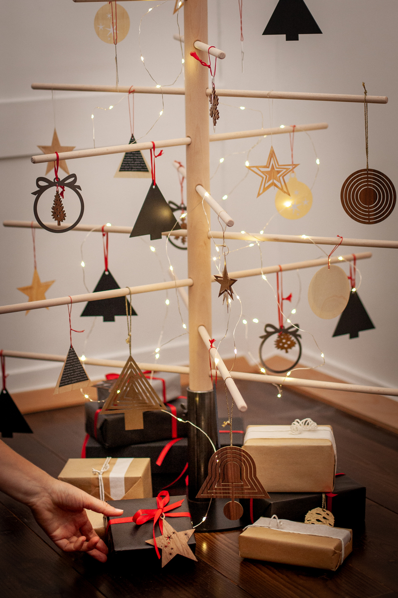 Ein abstrakter Weihnachtsbaum mit Stahlständer und dekorativen Holzornamenten steht in einem Innenraum. Der Baum ist mit Lichtern und verschiedenen Anhängern geschmückt, darunter Sterne und Kreise. Unter dem Baum befinden sich sorgfältig verpackte Geschenke in braunem und schwarzem Papier, verziert mit roten Schleifen.