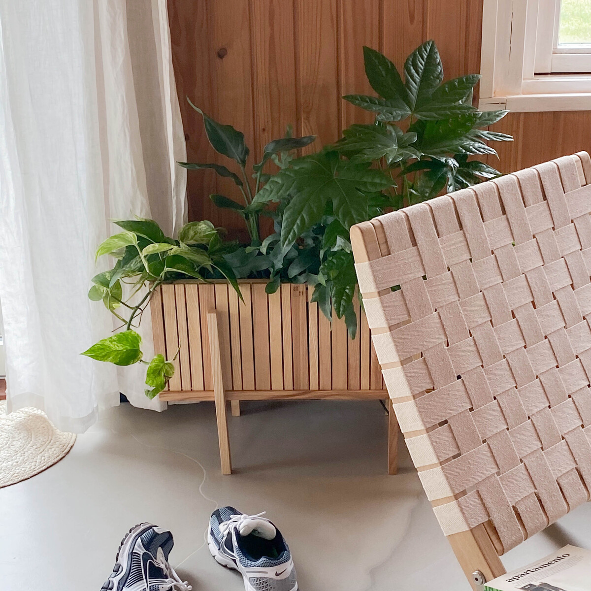 Pflanzgefäß »GrowWIDE« aus Holz mit grünen Zimmerpflanzen, minimalistischen Design und skandinavischem Stil. Im Vordergrund befindet sich ein moderner Lehnstuhl und ein Paar Sportschuhe auf einem hellgrauen Boden.