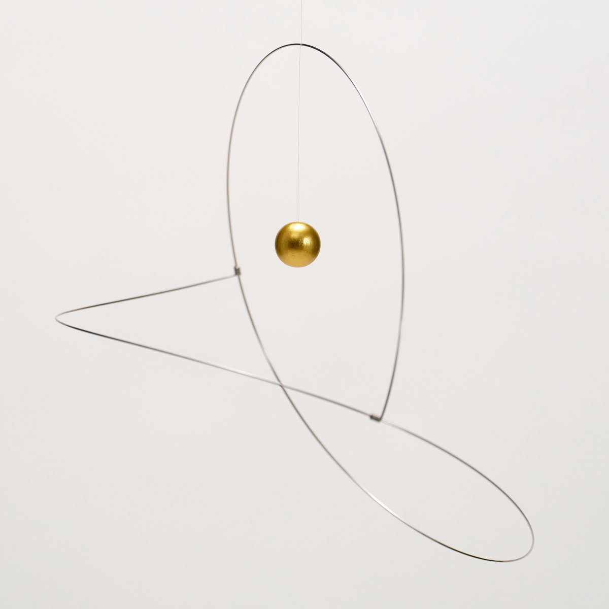 Das Bild zeigt das Mobile "Metamorphosis, Golden Planet" von Flensted Hanging Art. Eine goldene Kugel schwebt im Zentrum zweier eleganter, oval gebogener silberner Metalldrähte vor weißem Hintergrund. Das Design wirkt modern, leicht und minimalistisch.