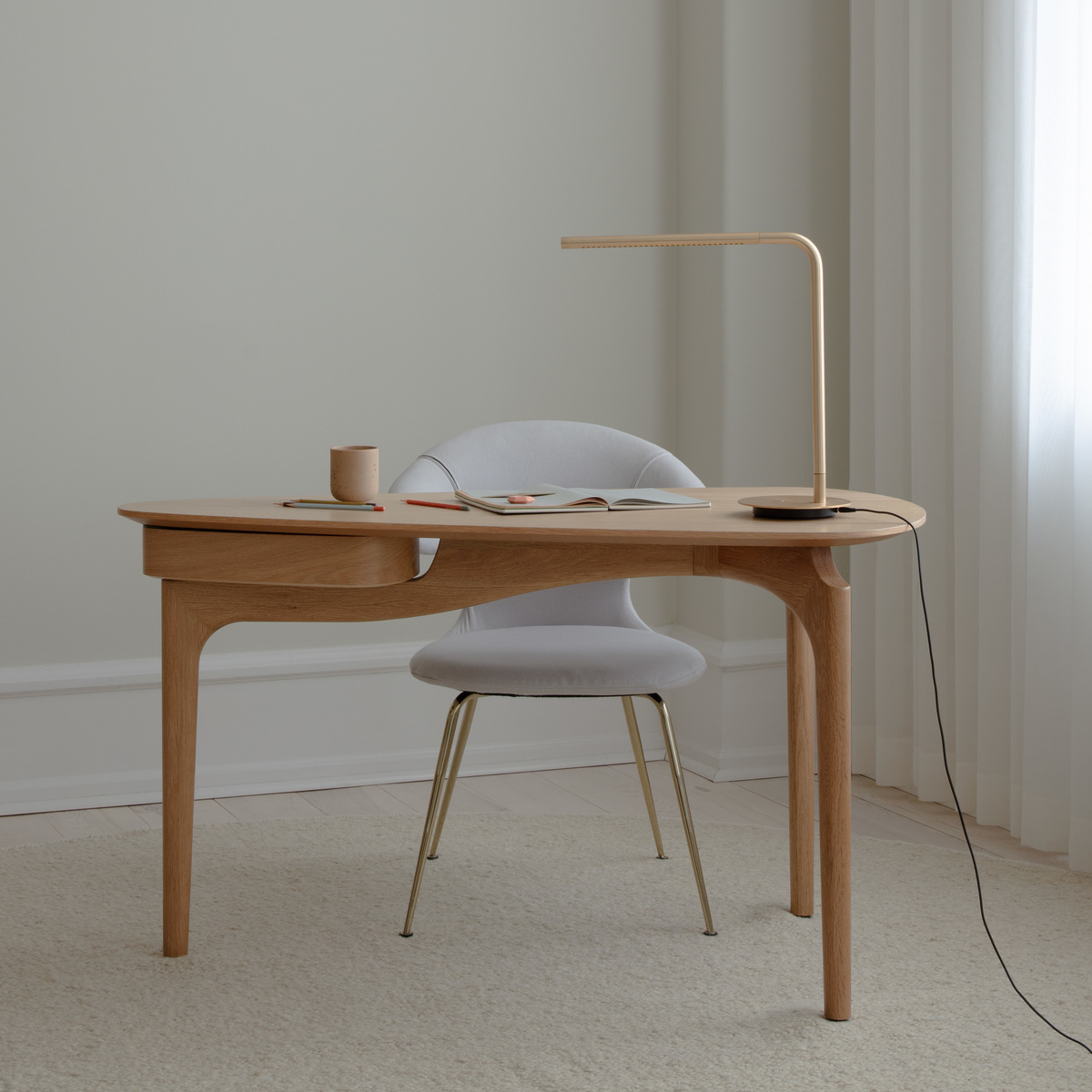 Moderner Schreibtisch aus hellem Holz mit abgerundeten Kanten steht vor einer hellen Wand und einem Fenster mit weißen Vorhängen. Auf dem Schreibtisch befinden sich eine minimalistische Lampe, ein Becher und Unterlagen. Ein gepolsterter Stuhl steht davor, alles in einer hellen, ruhigen Umgebung auf Teppichboden.