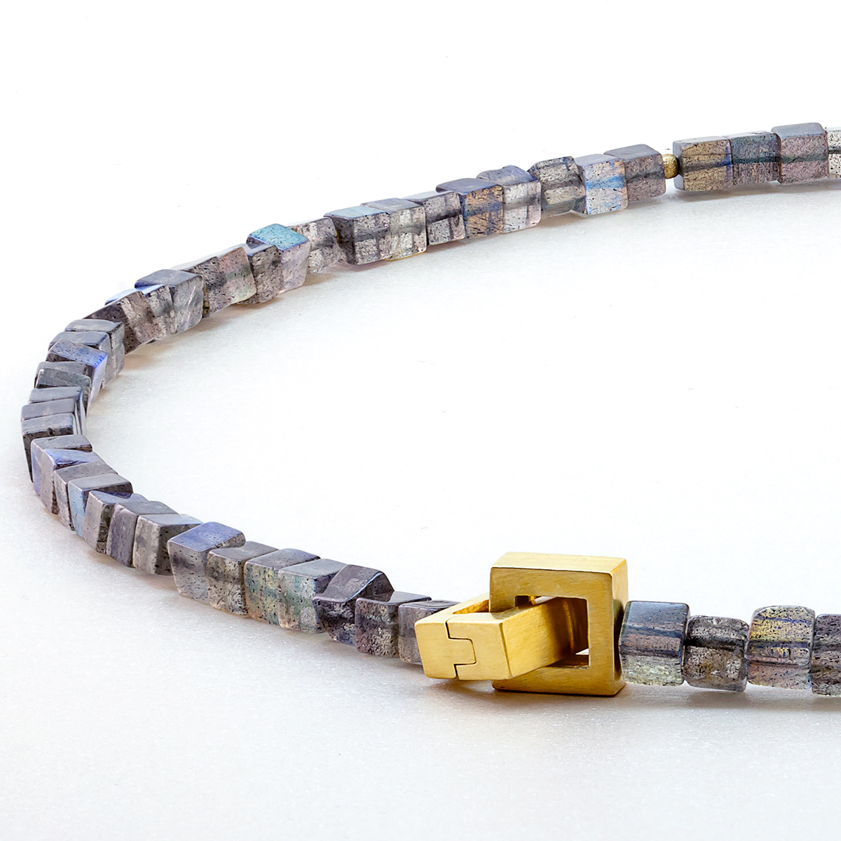 Abgebildet ist das Labradorit Collier von Menard. Die Kette besteht aus kleinen, kubischen Labradorit-Steinen mit irisierenden Grautönen, die von mattierten goldplattierten Silberkugeln unterbrochen werden. Mittelpunkt ist eine eckige, goldplattierte Patentschließe.