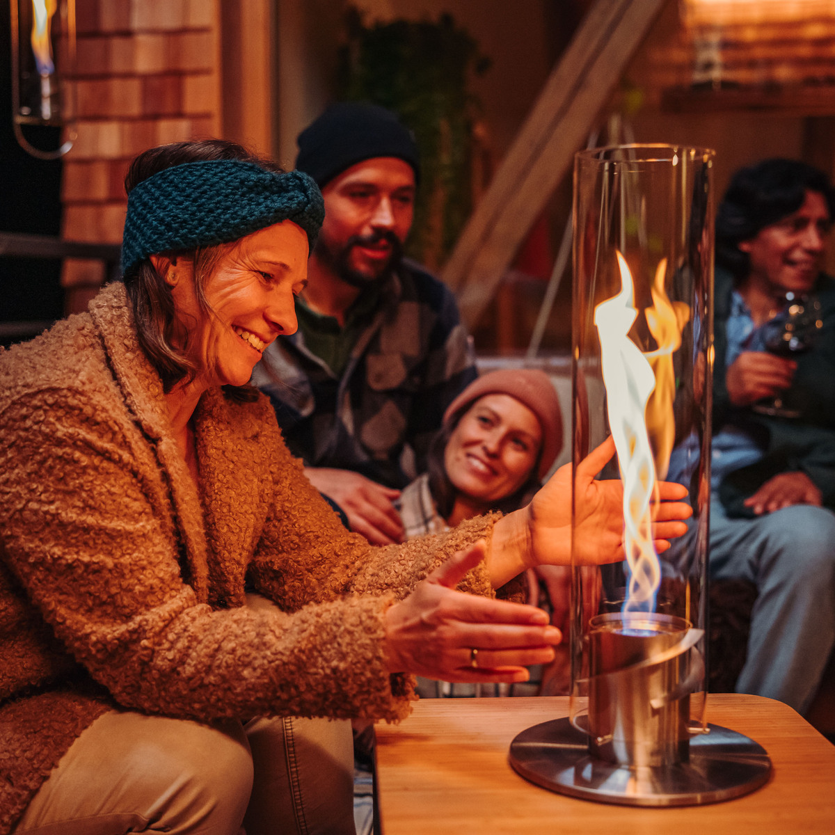 Vier Menschen sitzen in einer gemütlichen Umgebung. Eine Frau hält ihre Hände in die Nähe eines großen, zylindrischen Tischfeuer »SPIN 1200«, das auf einem Holztisch in der Mitte steht. Die Flammen sorgen für eine warme und einladende Atmosphäre.