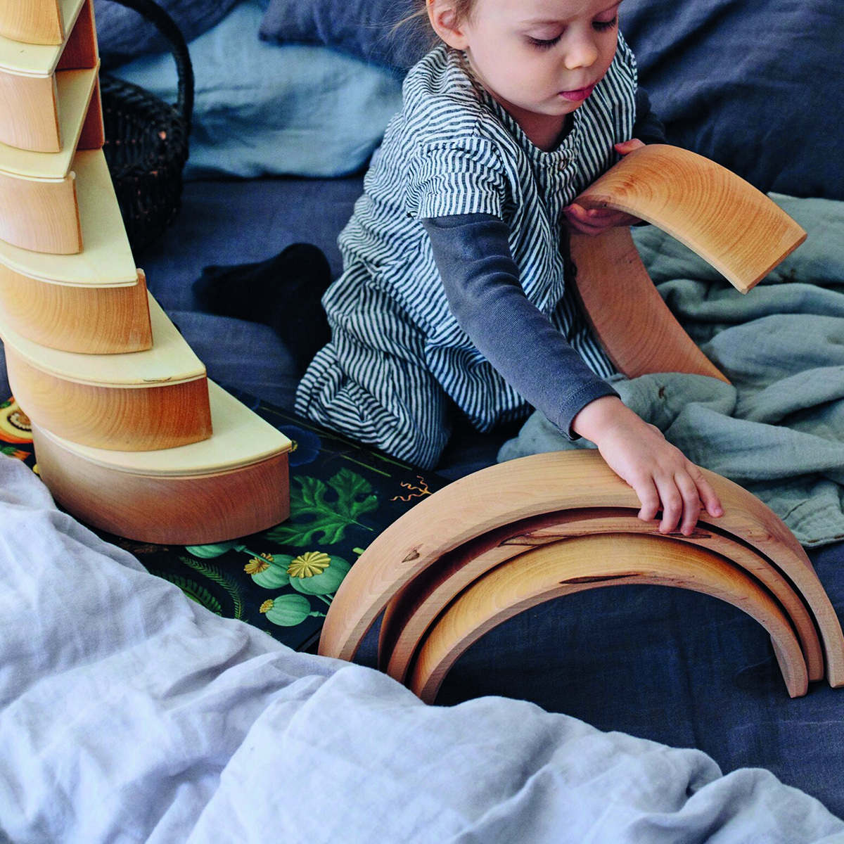Ein Kind spielt mit einem Regenbogen-Holzspielzeug auf einem Bett. Das Spielzeug besteht aus mehreren halbkreisförmigen Holzteilen, die zusammen einen Regenbogen bilden. Das Kind greift eines der Holzteile und ist in vertieftes Spiel vertieft. Im Hintergrund sind Decken und Kissen zu sehen. Das Holzspielzeug fördert die Kreativität und Motorik der Kinder.