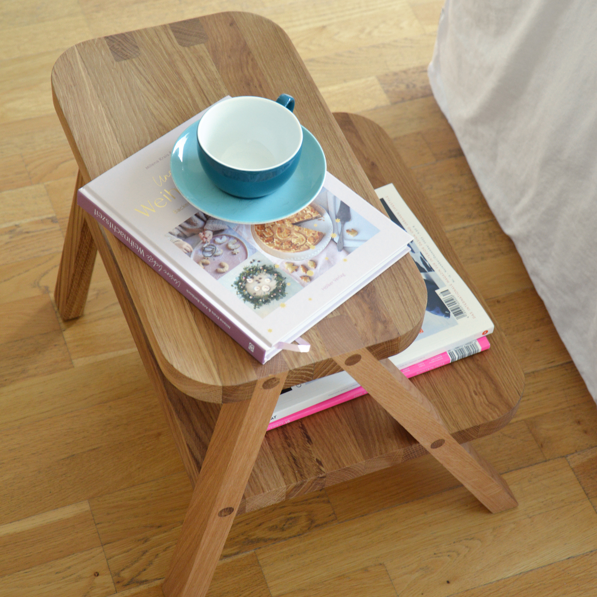 Ein zweistufiger Tritthocker aus hellem Holz, benutzt als Beistelltisch, mit einer Tasse und Büchern darauf. Der Hocker steht auf einem hellen Holzboden neben einem weißen Sofa.