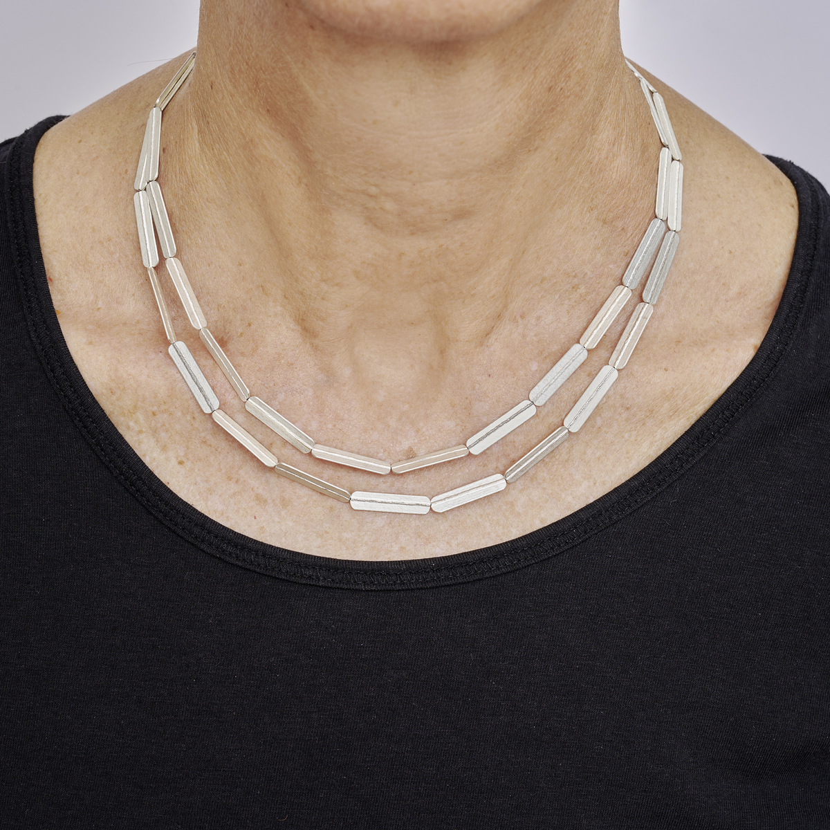Zweireihiges Collier aus geprägtem Silber mit Streifenmuster, getragen von einer erwachsenen Person in einem schwarzen Oberteil, abgebildet vor neutralem Hintergrund.