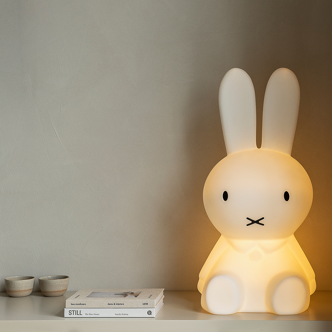 Das Bild zeigt eine »Miffy XL« Kinderlampe in Form eines großen, weißen Hasen, der auf einem Tisch neben zwei kleinen Schalen und einem Buch steht. Die Lampe leuchtet sanft und bietet eine beruhigende Beleuchtung, ideal für Kinderzimmer.