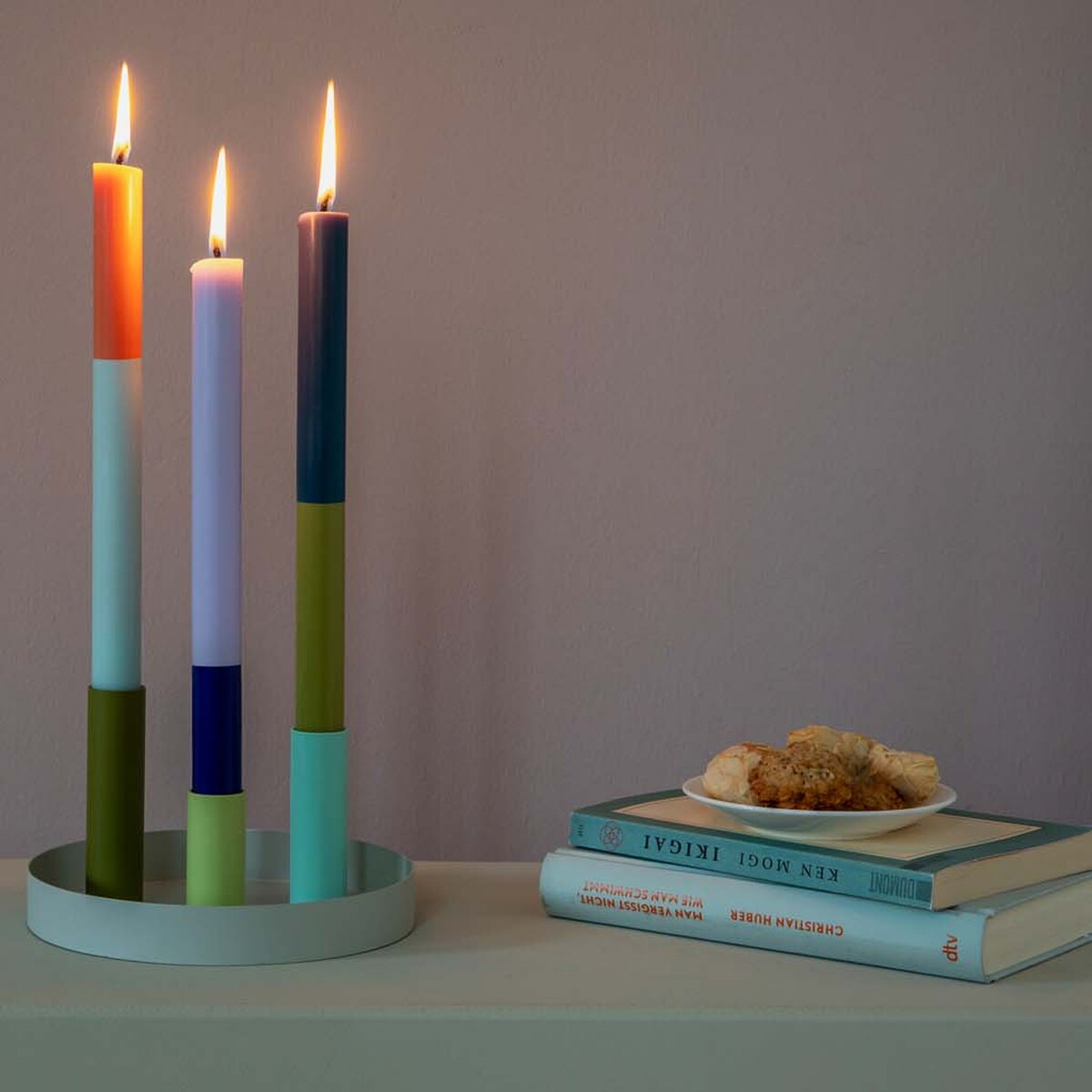Ein Set von drei brennenden Stabkerzen in unterschiedlichen Farben (Orange, Blau, Lila, Grün, Weiß) auf einem runden Kerzenhalter auf einem Tisch. Daneben liegen einige Bücher und ein kleiner Teller mit Gebäck.