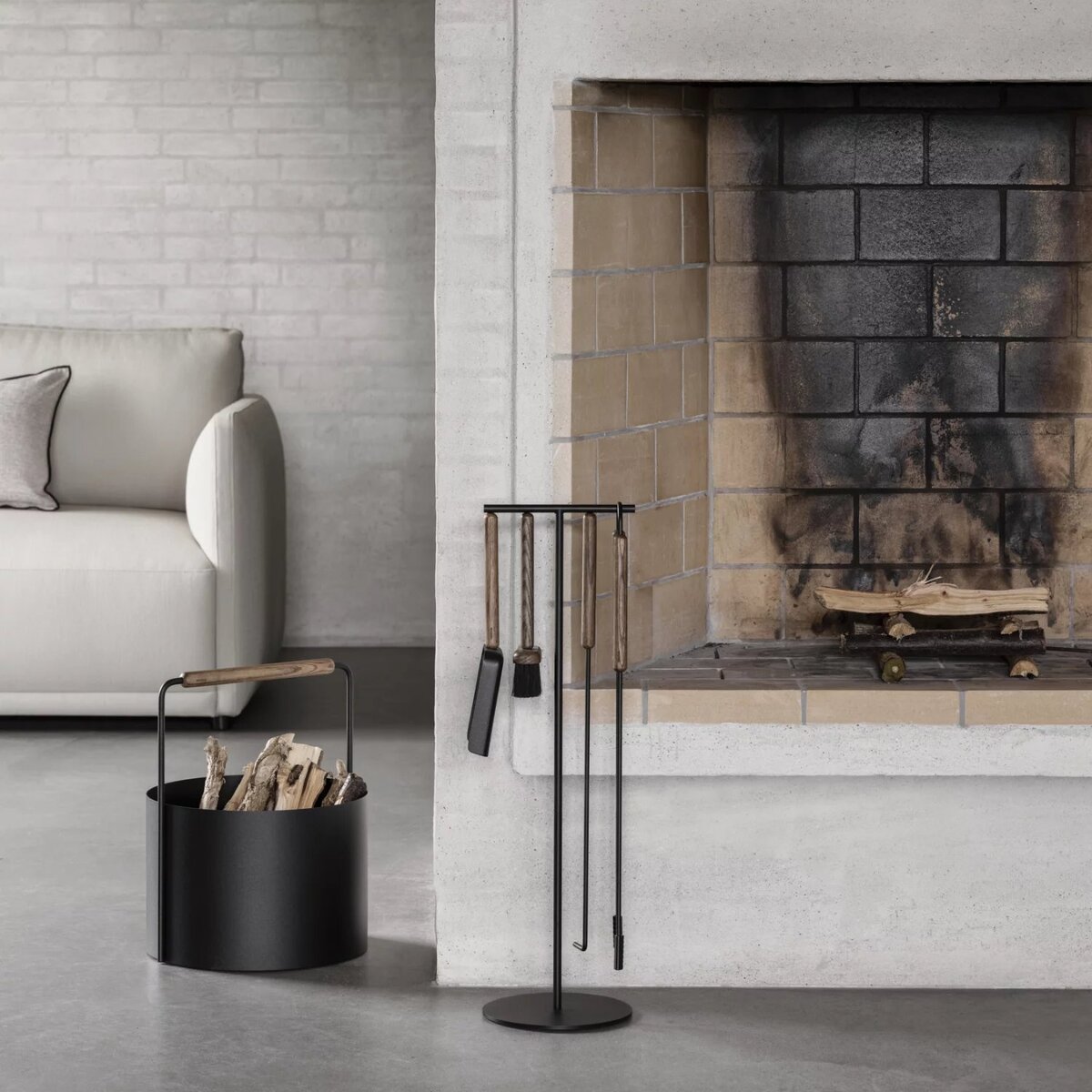 Moderner Holzkorb aus schwarzem Metall mit Holzhenkel steht neben einem offenen Kamin in einem minimalistisch eingerichteten Wohnzimmer mit heller Couch und grauen Wänden. Der Korb ist mit Feuerholz gefüllt und vermittelt ein wohnliches, skandinavisches Ambiente.