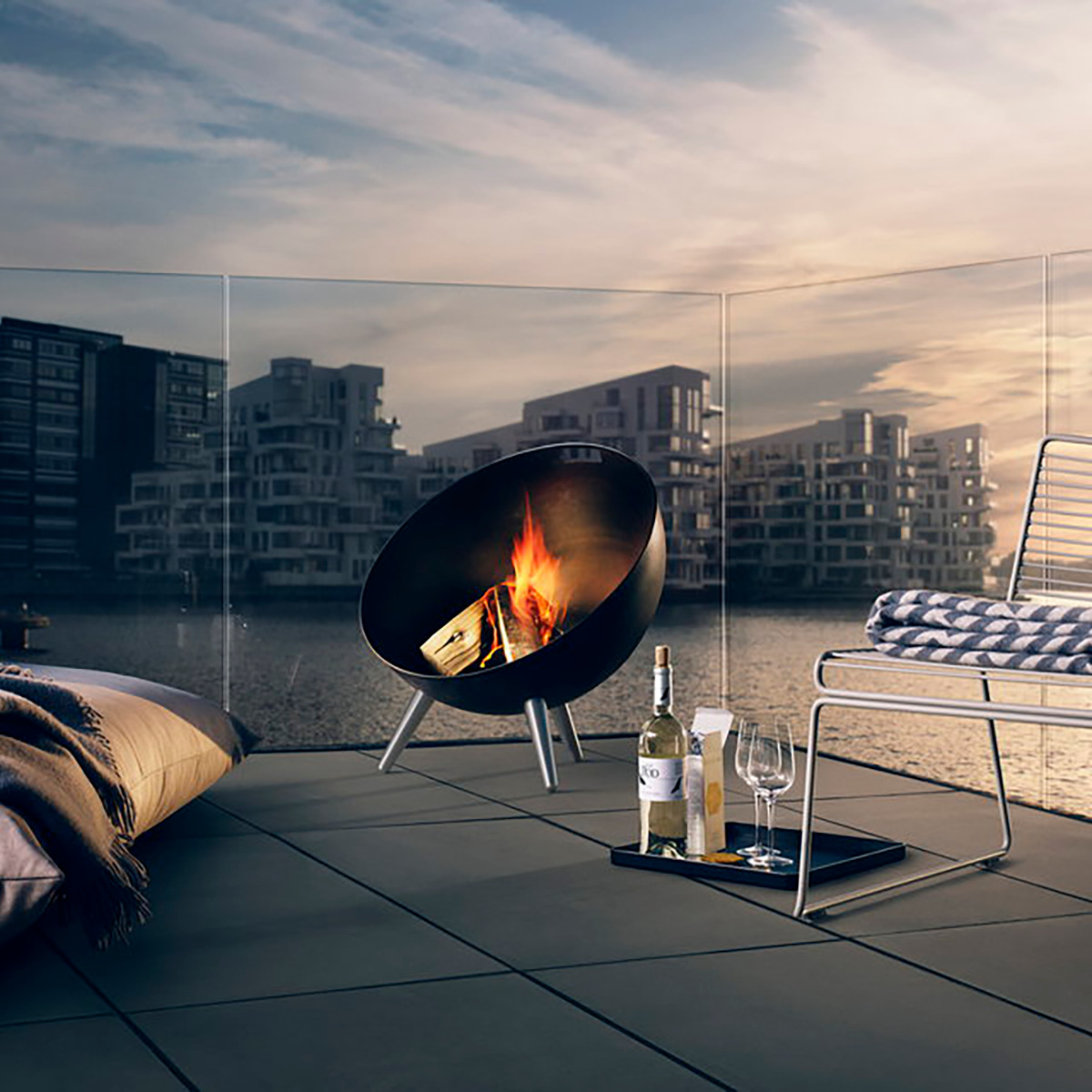 Ein stilvoller Außenbereich auf einer Terrasse bei Dämmerung, mit einer modernen Feuerschale namens »FireGlobe«, einem Hocker, einer Decke und einem Glas Wein daneben. Im Hintergrund sind moderne Wohngebäude zu sehen.