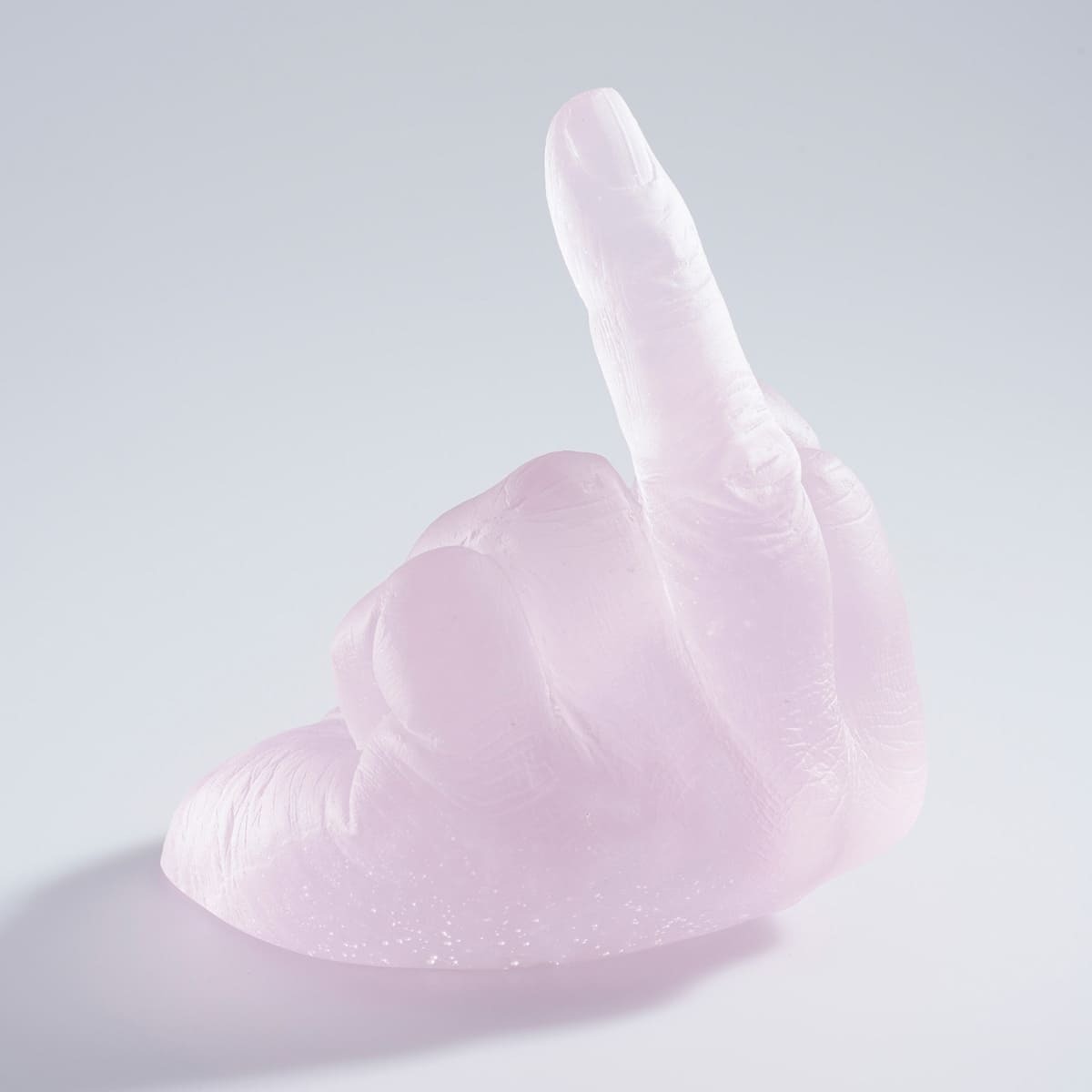 Eine Skulptur aus rosa transparentem Glas zeigt eine Hand, die den Mittelfinger hebt. Die Skulptur ist ein Kunstwerk von Ai Weiwei namens »Study of Perspective in Glass«. Sie liegt auf einer weißen, blanken Oberfläche.