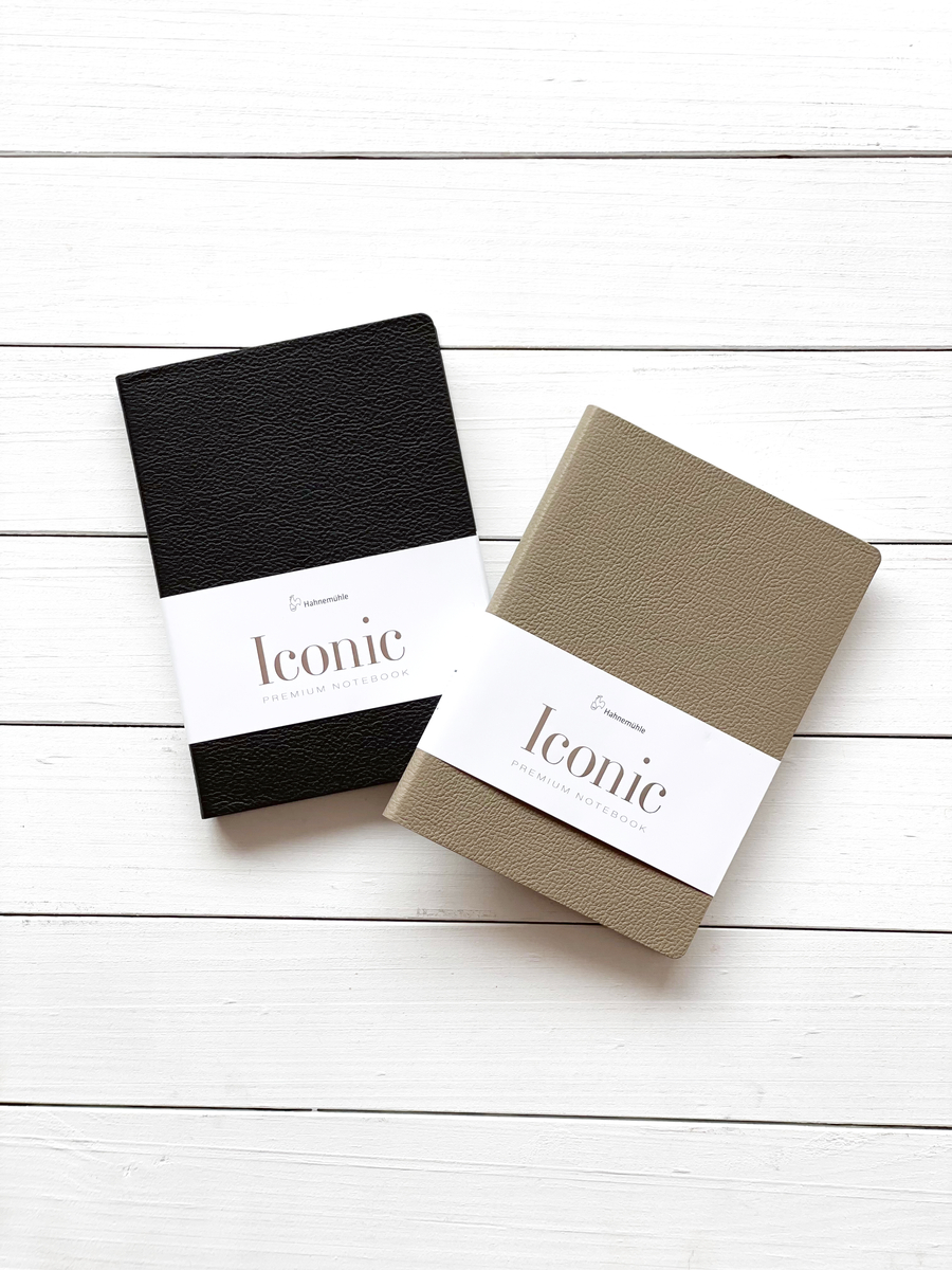 Zwei Notizbücher »Iconic« im DIN A5 Format auf einem weißen Holzuntergrund. Ein Notizbuch ist schwarz und das andere beige, beide werden von einer weißen Banderole umgeben.