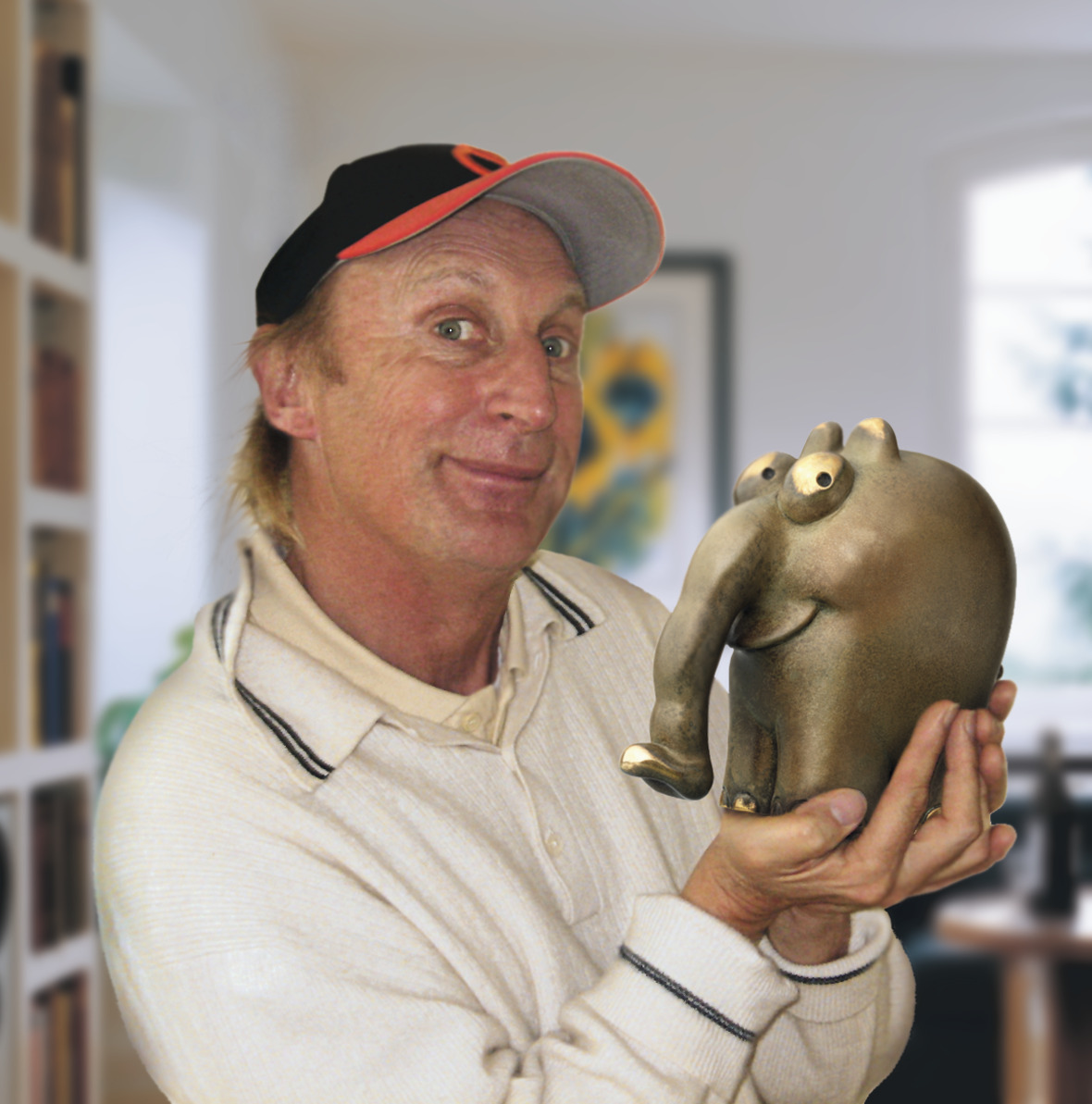 Otto Waalkes hält eine goldene Skulptur eines Ottifanten, lächelt und trägt eine schwarz-rote Baseballkappe sowie eine beige Jacke. Im Hintergrund sind raumhafte Details eines Wohnzimmers zu sehen.