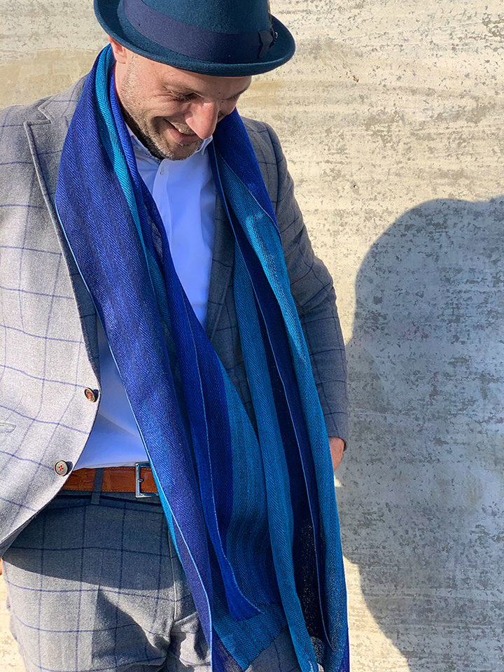 Ein Mann trägt einen breiten Schal aus blauem Leinen. Er hat einen eleganten Anzug an und steht vor einer Betonwand, die Sonnenlicht reflektiert. Der Schal hat verschiedene Blautöne und fällt locker über den Anzug.