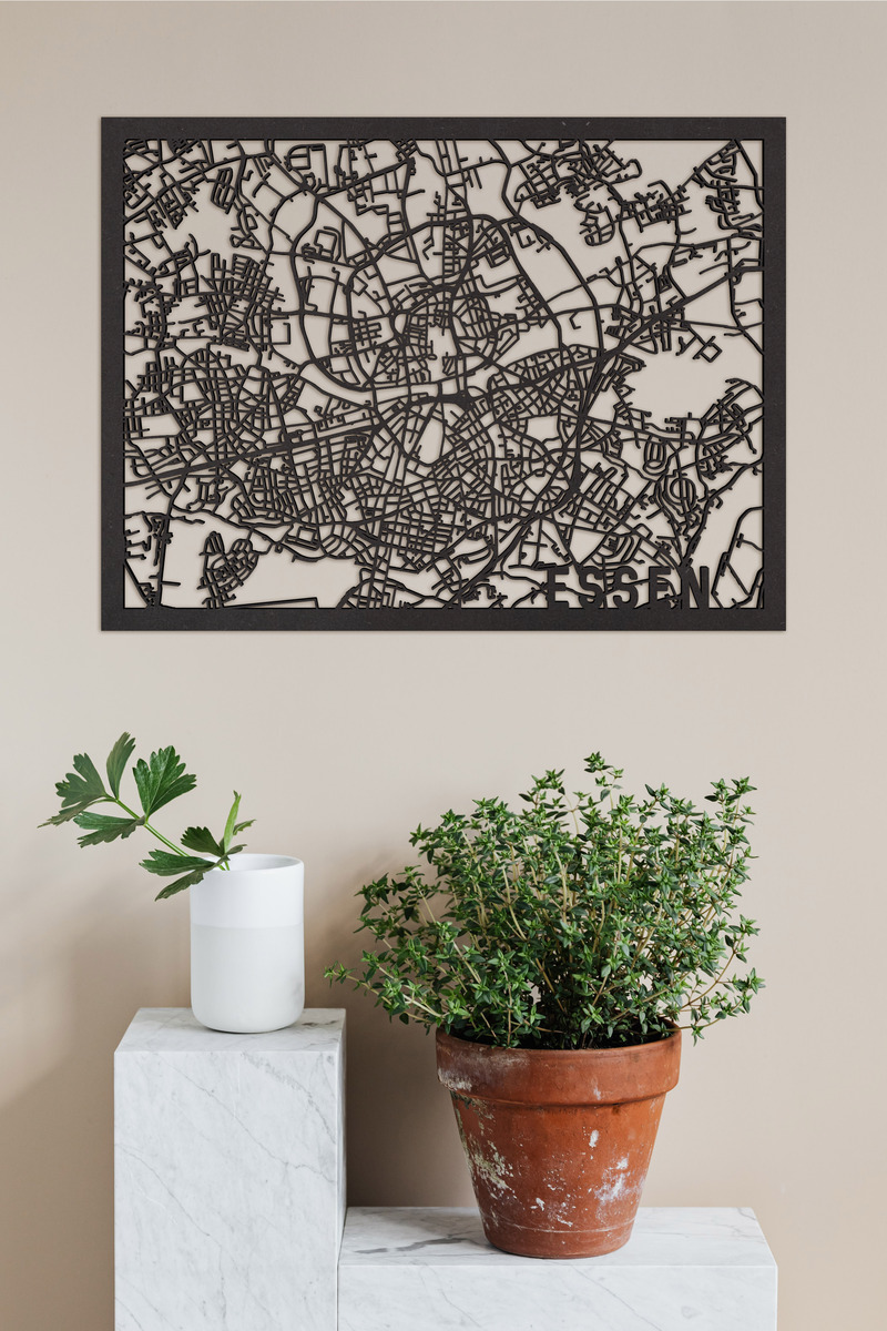 Eine detaillierte, schwarze Stadtkarte von Essen aus Holz, montiert auf einer weißen Wand. Vor der Wand stehen zwei Pflanzen, eine in einem Terrakottatopf und die andere in einem weißen Behälter, auf weißen, marmorähnlichen Blöcken.