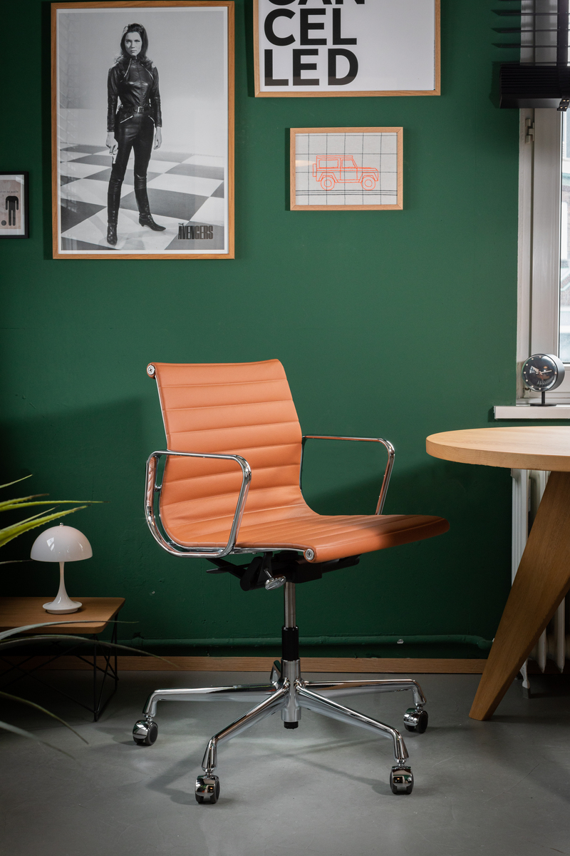 Der ZEIT-Sonderedition Vitra Alu Chair EA 118 ist ein verchromter Schreibtischstuhl mit brauner Polsterung. Er steht in einem modernen Büro vor einer grünen Wand, geschmückt mit Bilderrahmen. Eine Tischlampe, ein runder Holztisch und ein kleines Pflanzenelement sind ebenfalls sichtbar.