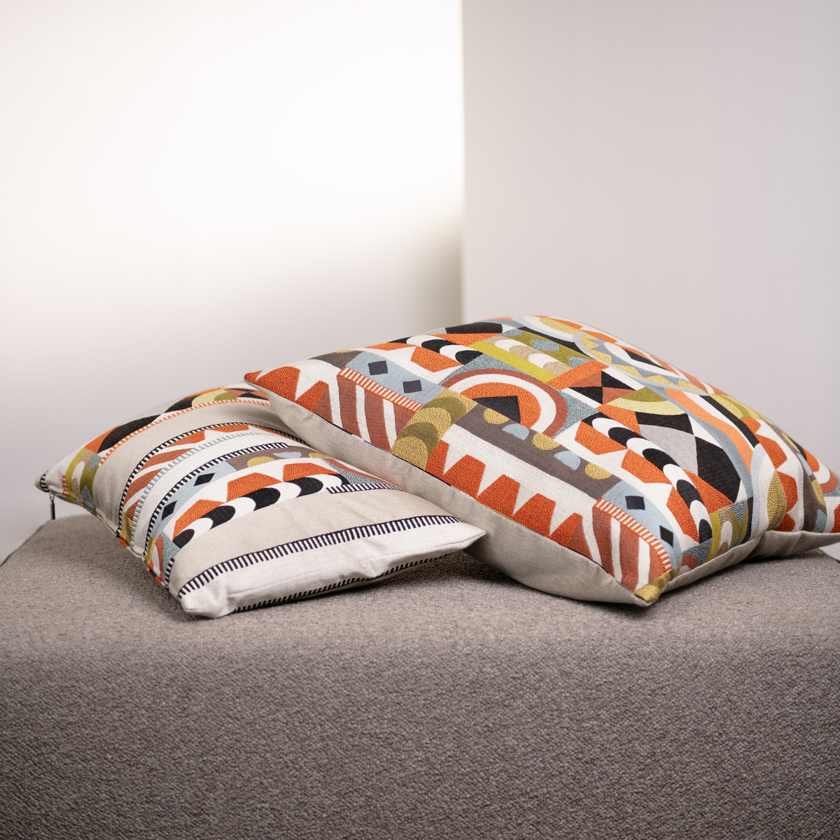 Zwei gemusterte Kissen im Havannah-Design liegen auf einem beigefarbenen Sofa. Die Kissen zeigen verschiedene geometrische Muster in Orange, Grün, Schwarz, Weiß und Beige.