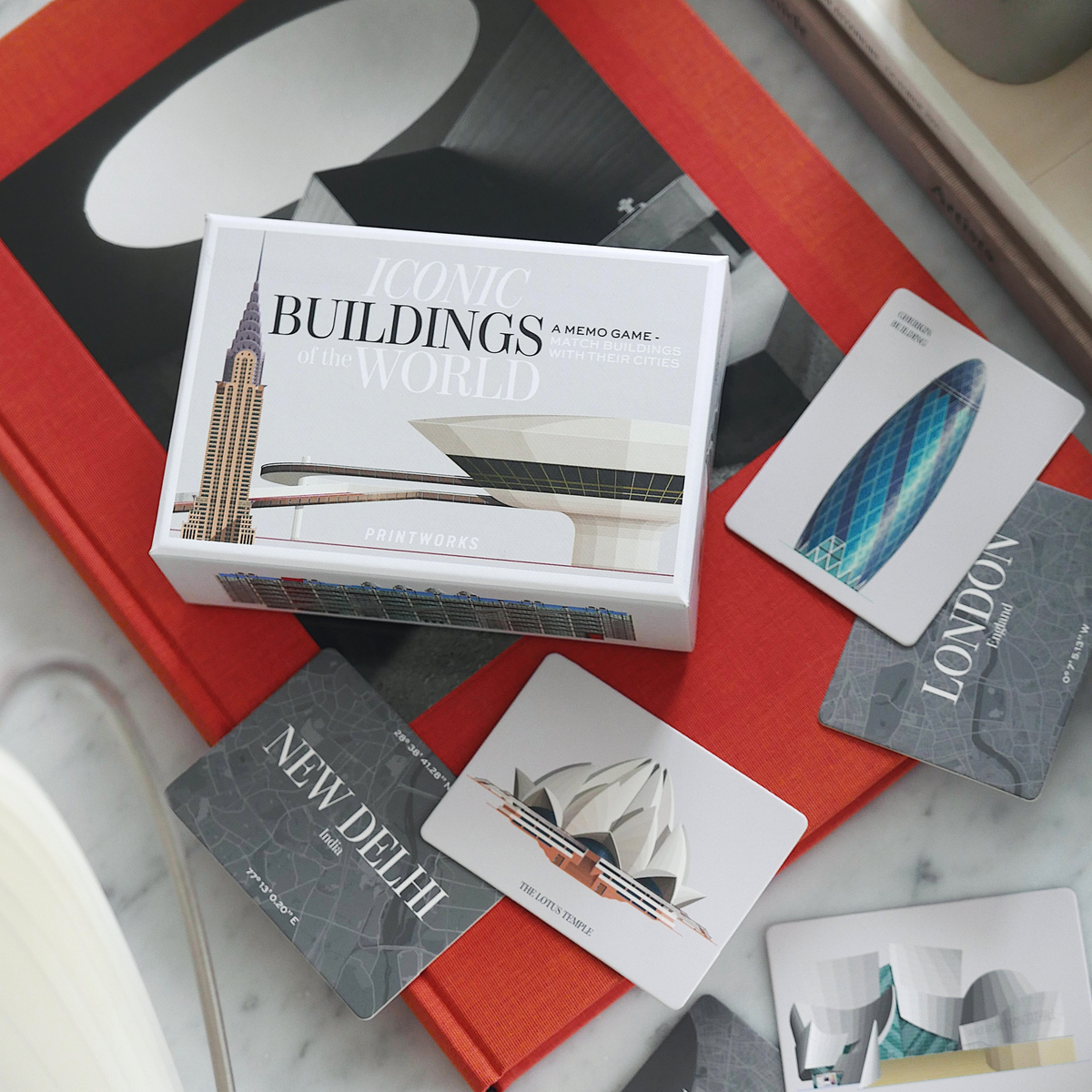 Das Gedächtnisspiel 'Architektur' zeigt verschiedene Spielkarten mit berühmten Bauwerken aus der ganzen Welt. Eine Spielbox mit der Aufschrift 'BUILDINGS of the WORLD' befindet sich im Zentrum. Zu den sichtbaren Karten gehören Wahrzeichen von New Delhi, London, Los Angeles und Sydney. Die Karten liegen auf einer weiß-marmorierten Tischplatte und einem roten Buch.