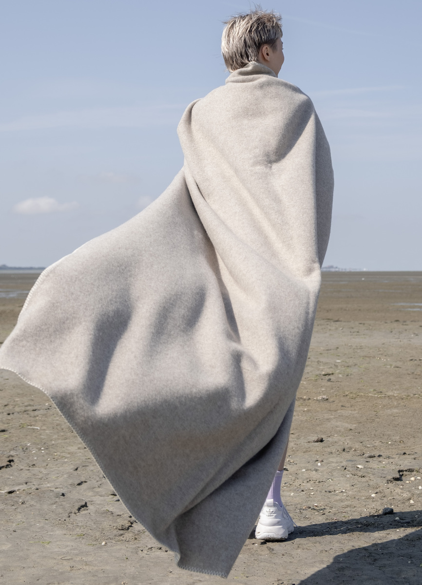 Eine Person steht am Strand und ist in eine große, hellgraue Merinodecke gehüllt. Die Decke weht leicht im Wind. Der Himmel ist blau und im Hintergrund ist Sand zu sehen. Die Szene vermittelt ein Gefühl von Ruhe und Entspannung.