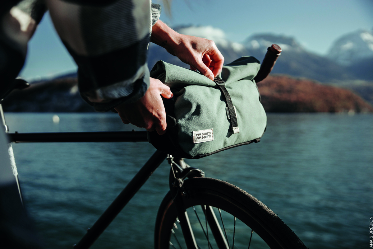 Person befestigt eine grüne Lenkertasche »Piha« an einem schwarzen Fahrradlenker, mit einem See und Bergen im Hintergrund.