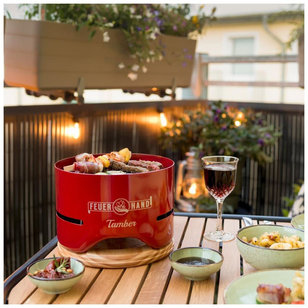 Ein roter Tischgrill »Tamber« von Feuerhand auf einem Holztisch auf einem Balkon, umgeben von Grünpflanzen und mehreren Tellern voller Essen. Im Hintergrund sind Lichter und Wein auf einem Glas zu sehen.