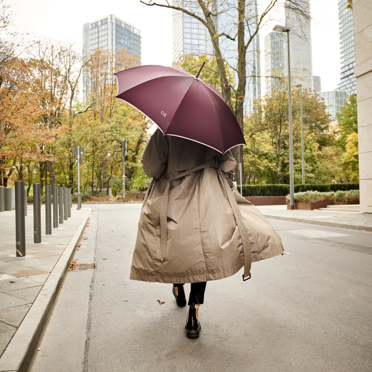 Eine Frau spaziert in einer beigen Trenchcoat bei herbstlichem Wetter durch eine städtische Umgebung und hält einen Beeren-farbenen Regenschirm 'Norfolk Oxford'. Hochhäuser und herbstliche Bäume sind im Hintergrund zu sehen.