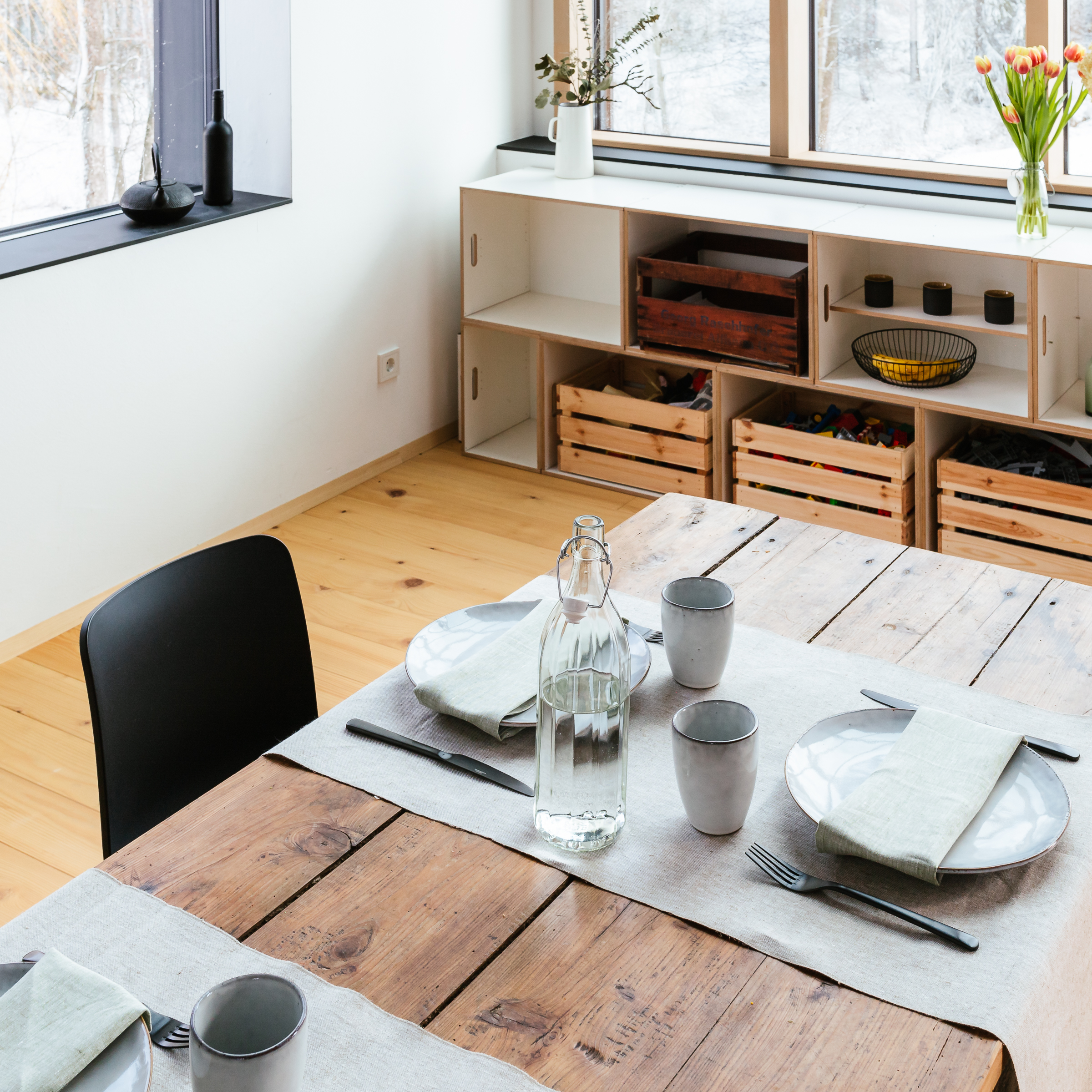 Leinen-Tischläufer liegt auf einem modernen Holztisch, gedeckt mit grauem Geschirr, Gläsern und einer klaren Wasserkaraffe in einem hell eingerichteten Esszimmer mit minimalistischer Dekoration.
