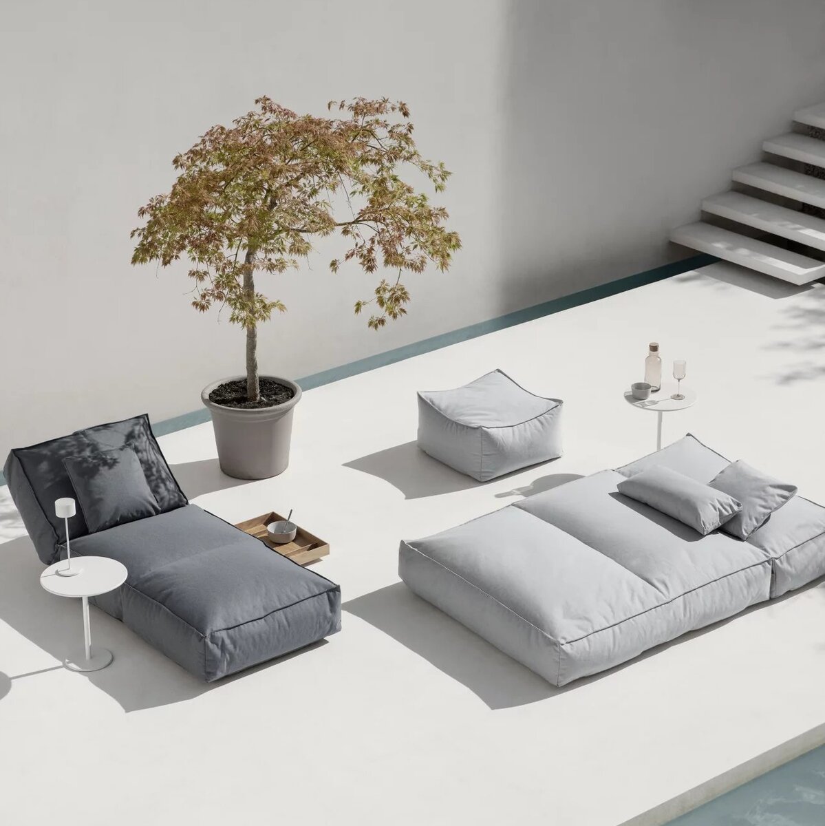 Auf dem Bild ist ein Outdoor-Bett »STAY« in den Maßen 80 x 190 cm zu sehen. Es steht auf einer weißen Terrasse neben einem kleinen runden Tisch, einem Lampenschirm und einem Blumenkübel mit einem Baum. Die Farben des Bildes sind hauptsächlich Grau, Weiß und Grün.
