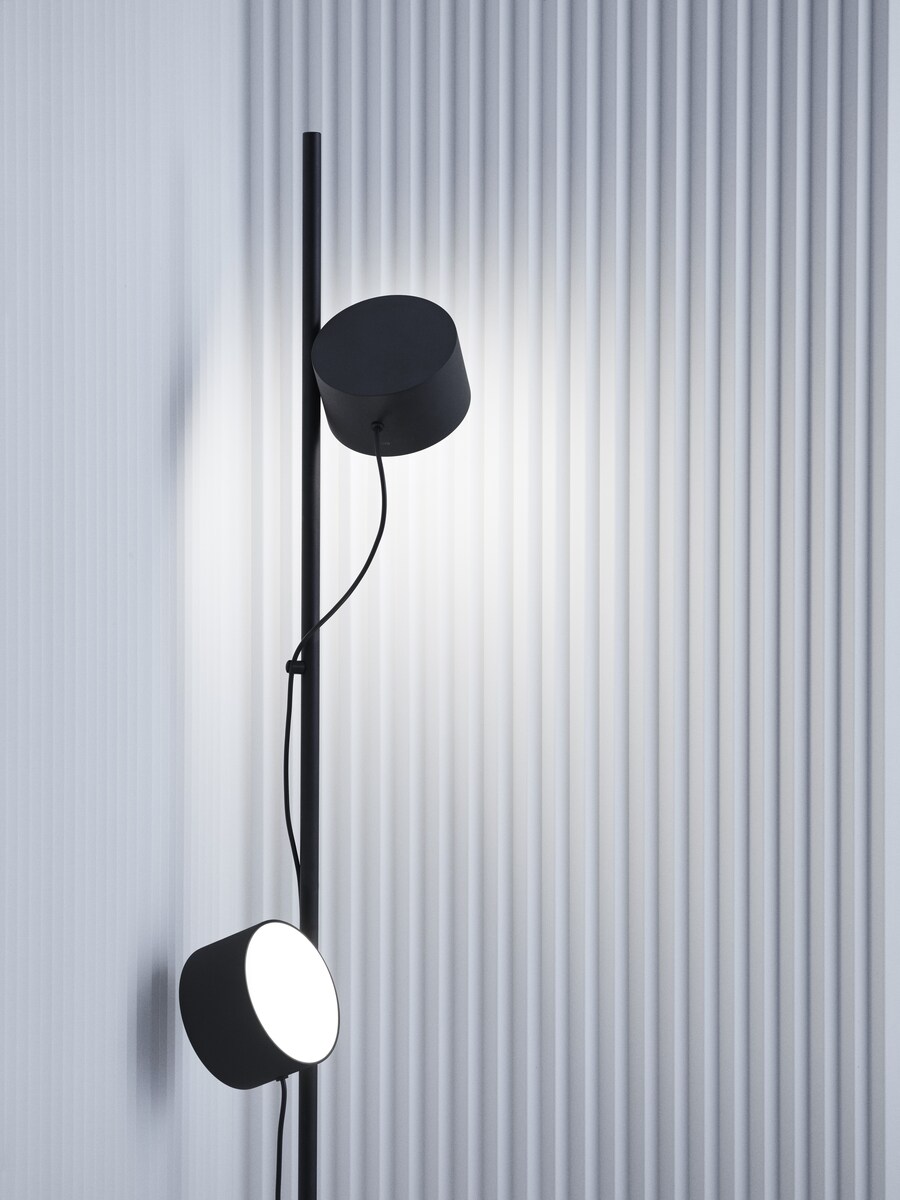 Moderne Bodenlampe »Post« mit minimalistischem Design. Die Lampe ist schwarz und hat zwei zylinderförmige Leuchten. Sie steht vor einer wellenförmigen, grauen Wand.