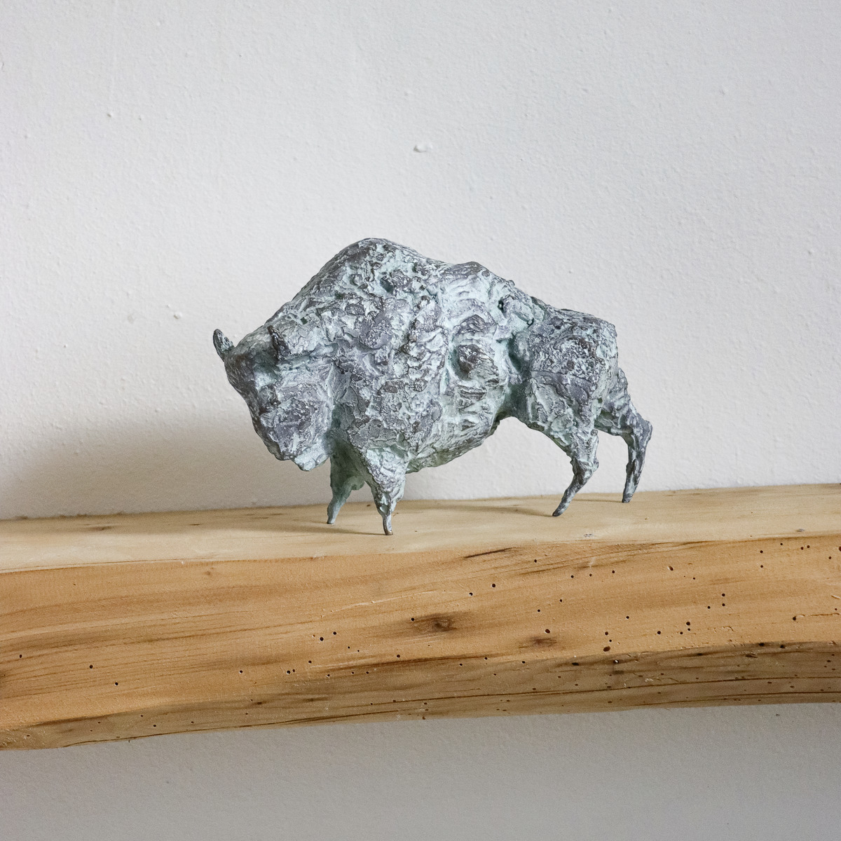 Bronzeskulptur eines Bisons von Michael Jastram auf einem naturbelassenen Holzbrett vor weißer Wand. Die Skulptur zeigt detailreiche Strukturen und einen grünlichen Patina-Effekt.