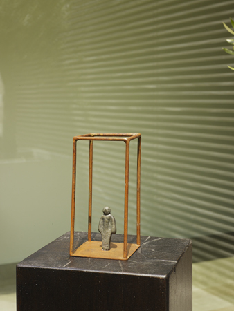 Ein Kunstwerk namens 'The Window Room' von Erli Fantini. Die Skulptur zeigt eine kleine Figur innerhalb eines rechteckigen Rahmens aus Metall auf einem schwarzen Sockel. Der Hintergrund ist grünlich und texturiert.