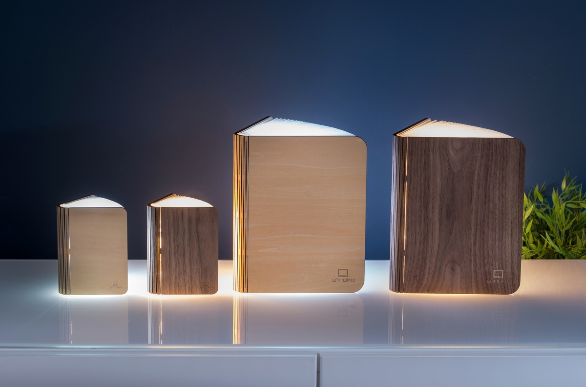 Das Bild zeigt vier Smart-Booklights »Ahorn« in verschiedenen Größen. Diese sind in Holzoptik gehalten und strahlen ein warmes, weißes Licht aus. Die Lampen sind wie Bücher gestaltet und sind auf einer weißen Oberfläche vor einem dunkelblauen Hintergrund platziert. Rechts im Bild ist eine kleine grüne Pflanze zu sehen.