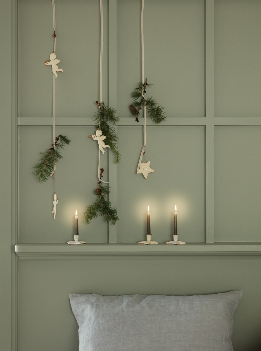 Ein Set von Weihnachtsanhängern namens 'Heilige Nacht', dekoriert mit Engeln und Sternen aus naturfarbenem Material, hängt an einer grünen Wand, geschmückt mit Tannenzweigen. Darunter stehen drei brennende Kerzen in schlichten Haltern.