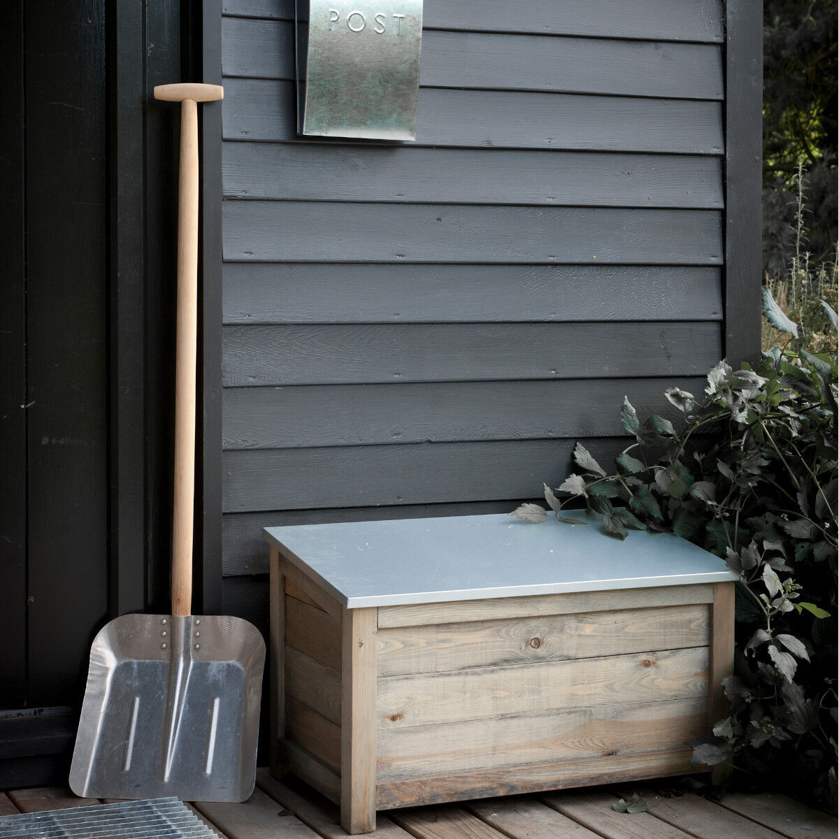Bild einer rustikalen Holz-Gartenbox »Aldsworth« neben einer großen Metallschaufel und einem silbernen Briefkasten an einer grau gestrichenen Wand. Die Kiste hat einen Deckel in helleren Farben. Neben der Holzbox sind grüne Pflanzen zu sehen.