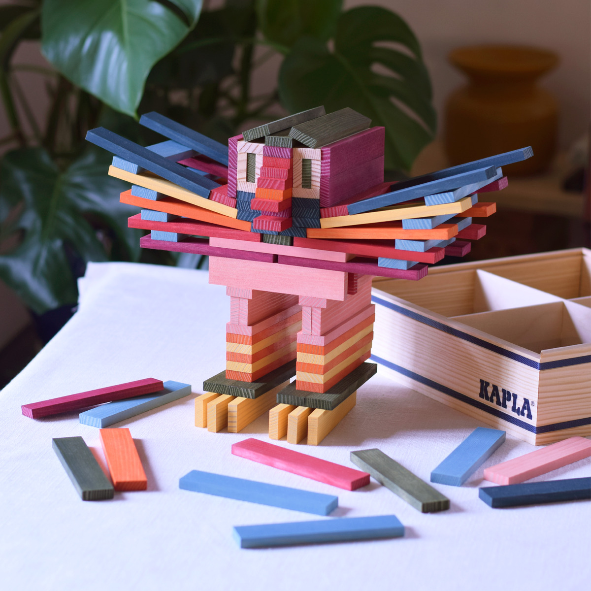 Ein Satz bunter Holzbausteine im 100er Kasten ist abgebildet. Verschiedene farbige Bausteine sind zu einer kreativen Figur zusammengesetzt, umgeben von weiteren Bausteinen auf einem weißen Untergrund. Im Hintergrund sind eine Zimmerpflanze und ein gelber Topf zu sehen.