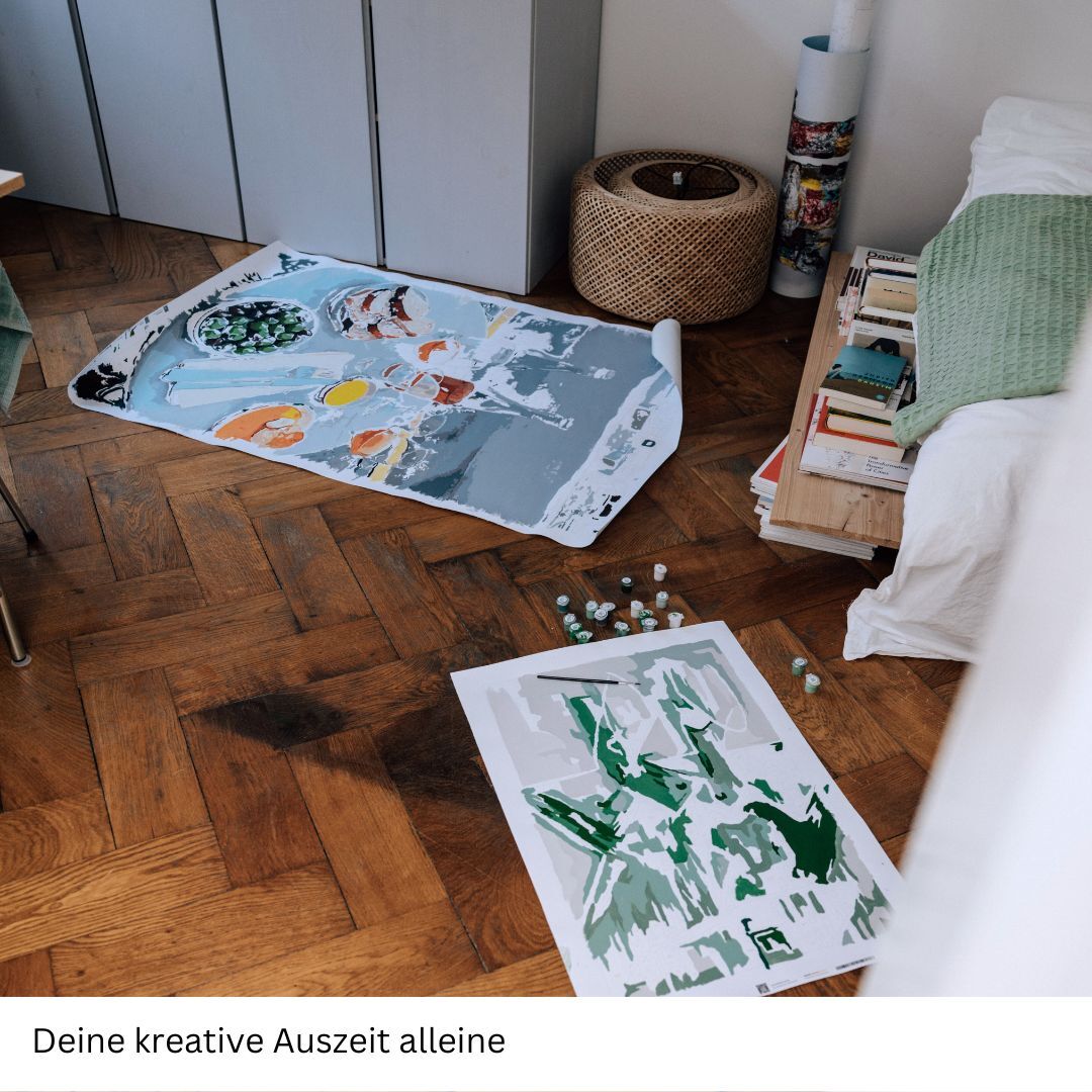 Ein Malen nach Zahlen Set mit dem Titel 'Schmeckt Oysters Gut' liegt auf einem Holzboden in einem hellen Zimmer. Die Leinwand zeigt teilweise gemalte Austermotive in grünen und blauen Tönen. Daneben liegen grüne und weiße Farbtöpfe und Pinsel. Im Hintergrund sind ein Korb, ein kleiner Schrank, Bücher und ein grünes Kissen auf einem Bett.