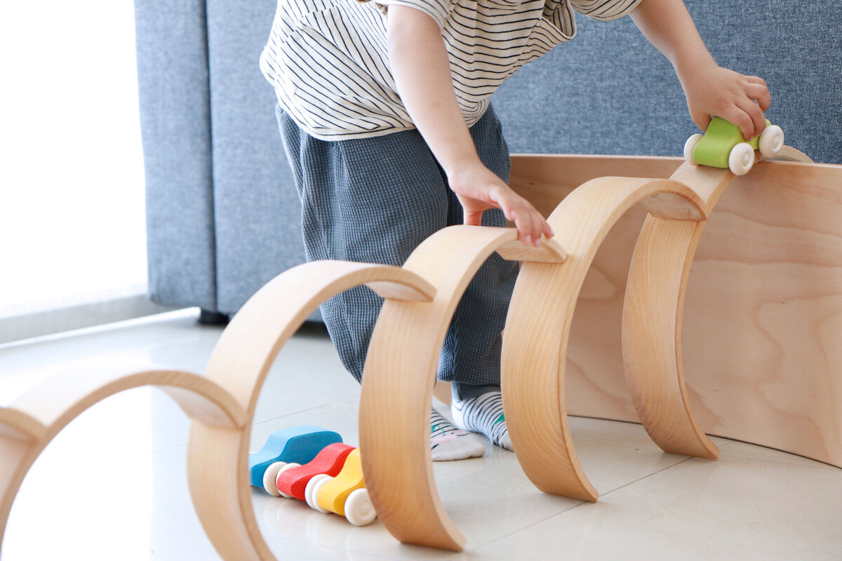 Ein Kind spielt mit einem Regenbogen Holzspielzeug. Das Spielzeug besteht aus natürlich gehaltenen, halbkreisförmigen Holzelementen, die übereinander gestapelt werden. Im Vordergrund liegen zusätzlich bunte, kleine Holzteile. Der Boden ist hell gefliest und im Hintergrund ist ein graues Sofa zu sehen.