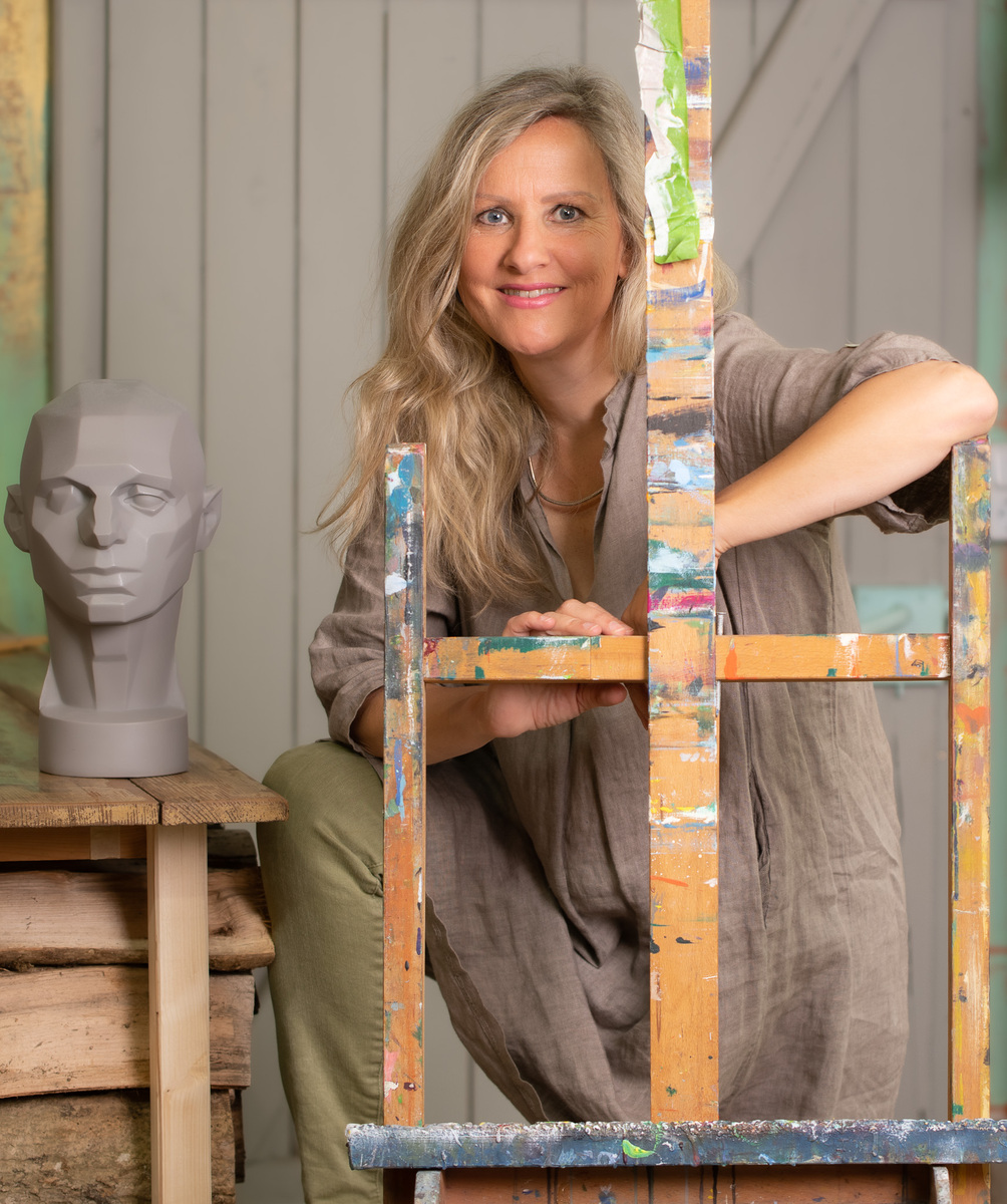Die Künstlerin Insa Hoffmann steht in ihrem Atelier neben einer bemalten Staffelei. Auf einem Tisch in der Nähe befindet sich eine graue Büste. Die Szene ist in natürlichen Farben wie Beige, Grau und Hellgrün gehalten.