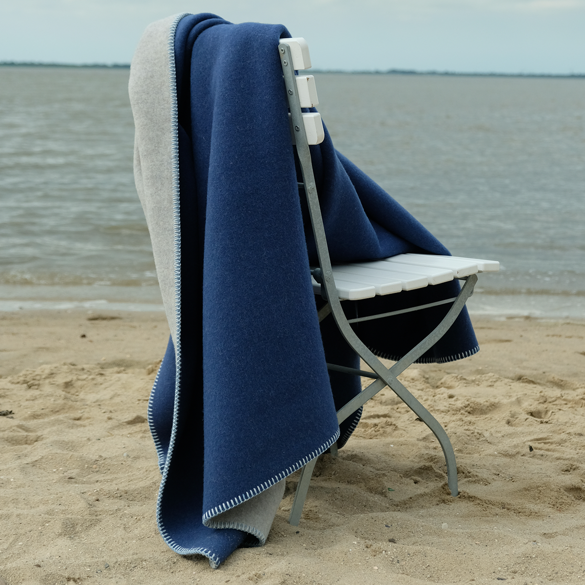 Eine Merino-Decke in Blau und Grau ist locker über einen weißen Klappstuhl gelegt, der am sandigen Ufer eines ruhigen Gewässers steht. Die Szene vermittelt eine entspannte, maritime Atmosphäre.