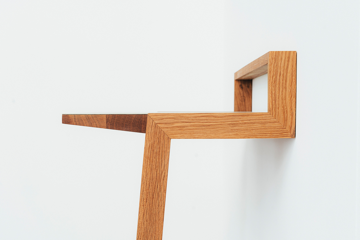 Das Bild zeigt ein minimalistisches Stehpult aus Holz, das an einer weißen Wand befestigt ist. Das Produkt hat ein schlichtes, modernes Design mit klaren Linien und eine natürliche, braune Holzfarbe.
