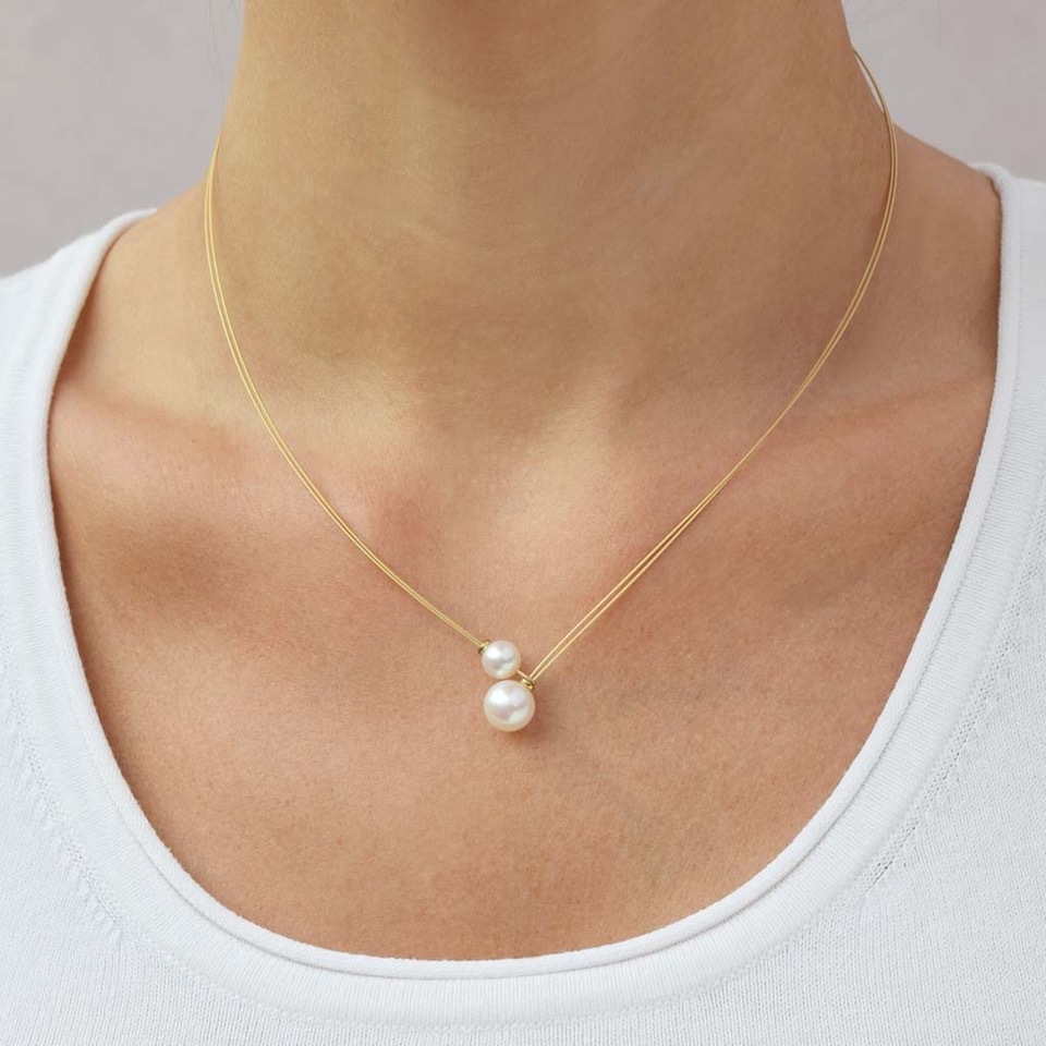 Eine schlanke goldfarbene Halskette mit zwei weißen Perlen, die nebeneinander als Anhänger arrangiert sind. Die Kette liegt elegant am Hals einer Frau, die ein weißes Oberteil trägt. Der Hintergrund ist neutral gehalten.