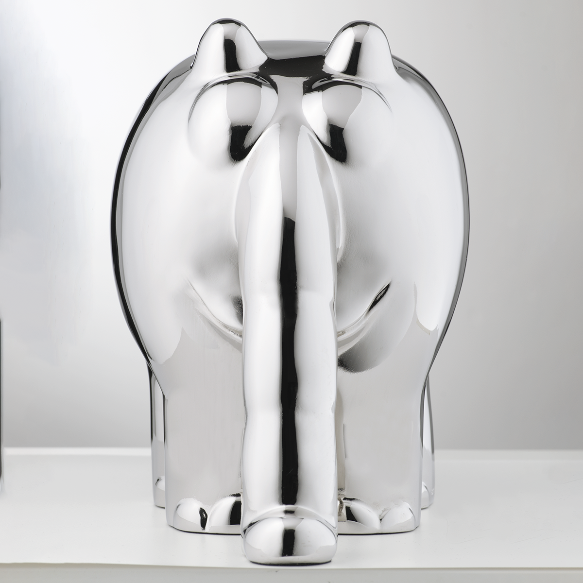 Die Skulptur zeigt einen stilisierten Ottifanten in glänzendem Metallic-Silber. Die Figur steht aufrecht auf einer glatten, weißen Fläche und ist rundlich geformt. Erkennbar sind charakteristische Formen wie der lange Rüssel und die großen Ohren. Das Licht reflektiert die glatte Oberfläche.