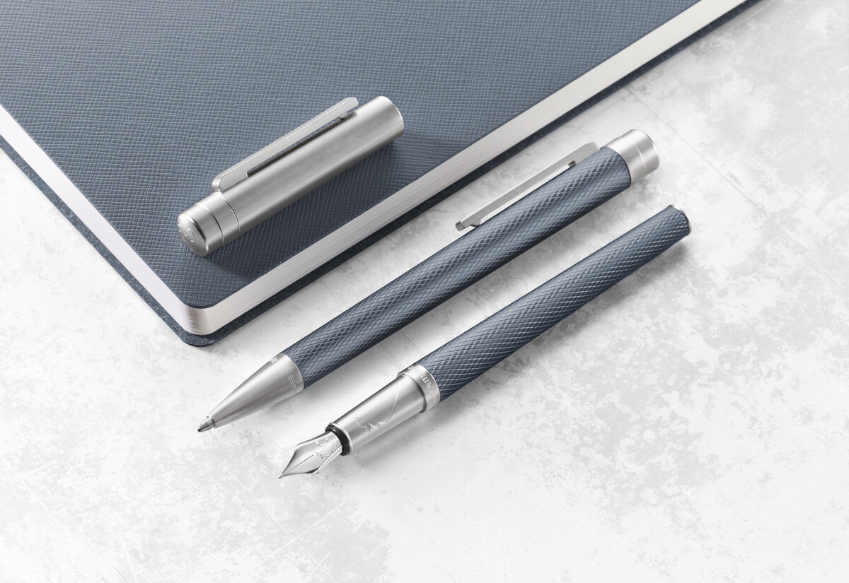 Ein Füllfederhalter der Slim Edition in Blau und Silber, neben einem passenden Notizbuch, auf einer hellen Oberfläche liegend.