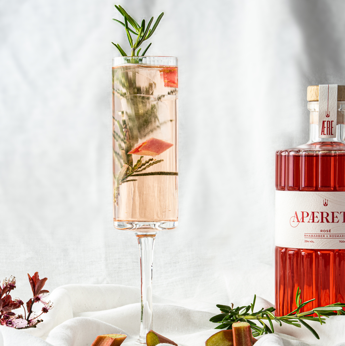 Ein Glas AERE Aperitif »Rosé« steht auf einem weißen Leinentuch, umgeben von Rosmarinzweigen und Rhabarberstücken. Im Hintergrund ist eine dazugehörige Flasche des Aperitifs zu sehen.