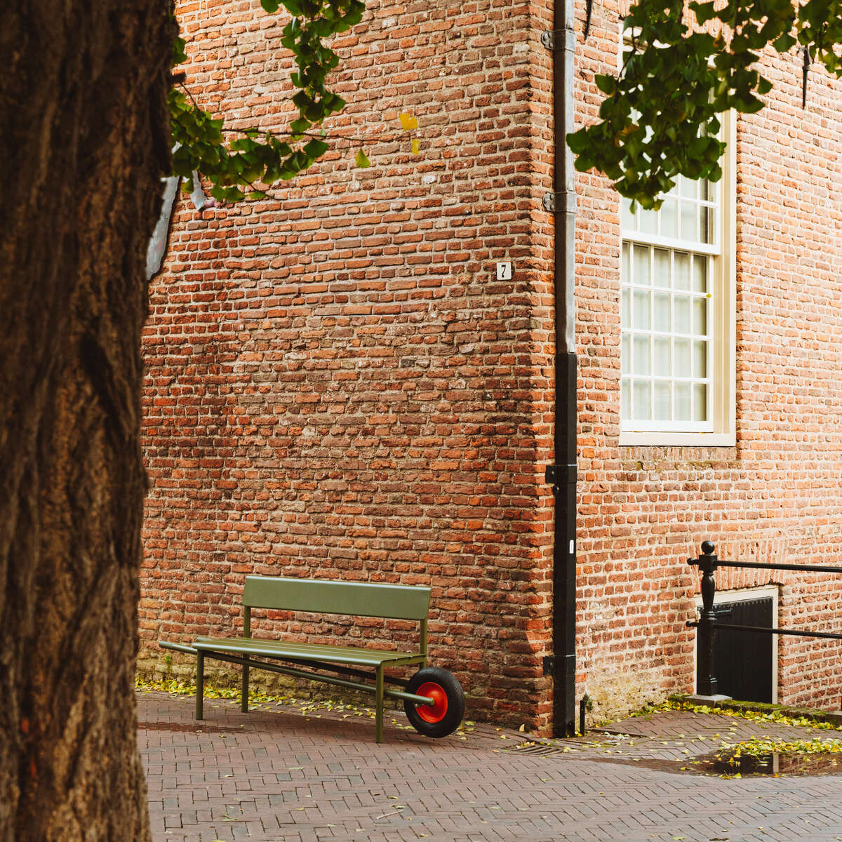 Eine Schubkarrenbank »Wheelbench« von Weltevree steht auf einem gepflasterten Platz vor einer roten Backsteinwand, flankiert von einem alten Baum. Die Bank hat ein grünes Sitzteil und ein markantes rotes Rad, umgeben von herabfallenden Blättern und urbaner Atmosphäre.