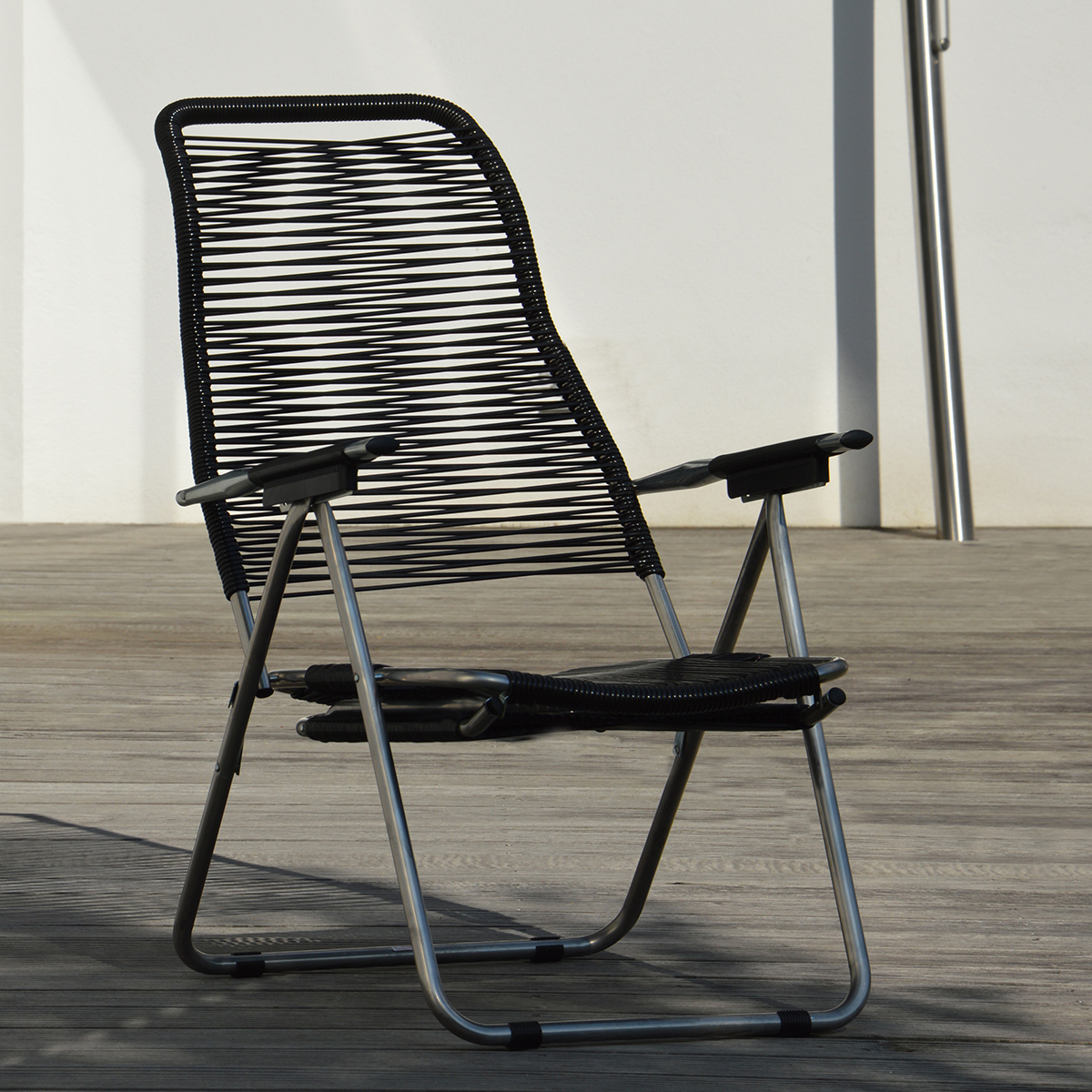 Schwarzer Armlehnsessel »Spaghetti« von Jan Kurtz steht auf einer Terrasse mit Holzplanken vor weißer Wand. Das Gestell ist aus silberfarbenem Aluminium, die Sitzfläche und Rückenlehne bestehen aus schwarz gespannter PVC-Schnürung im 80er Jahre-Design. Modern, filigran und komfortabel.