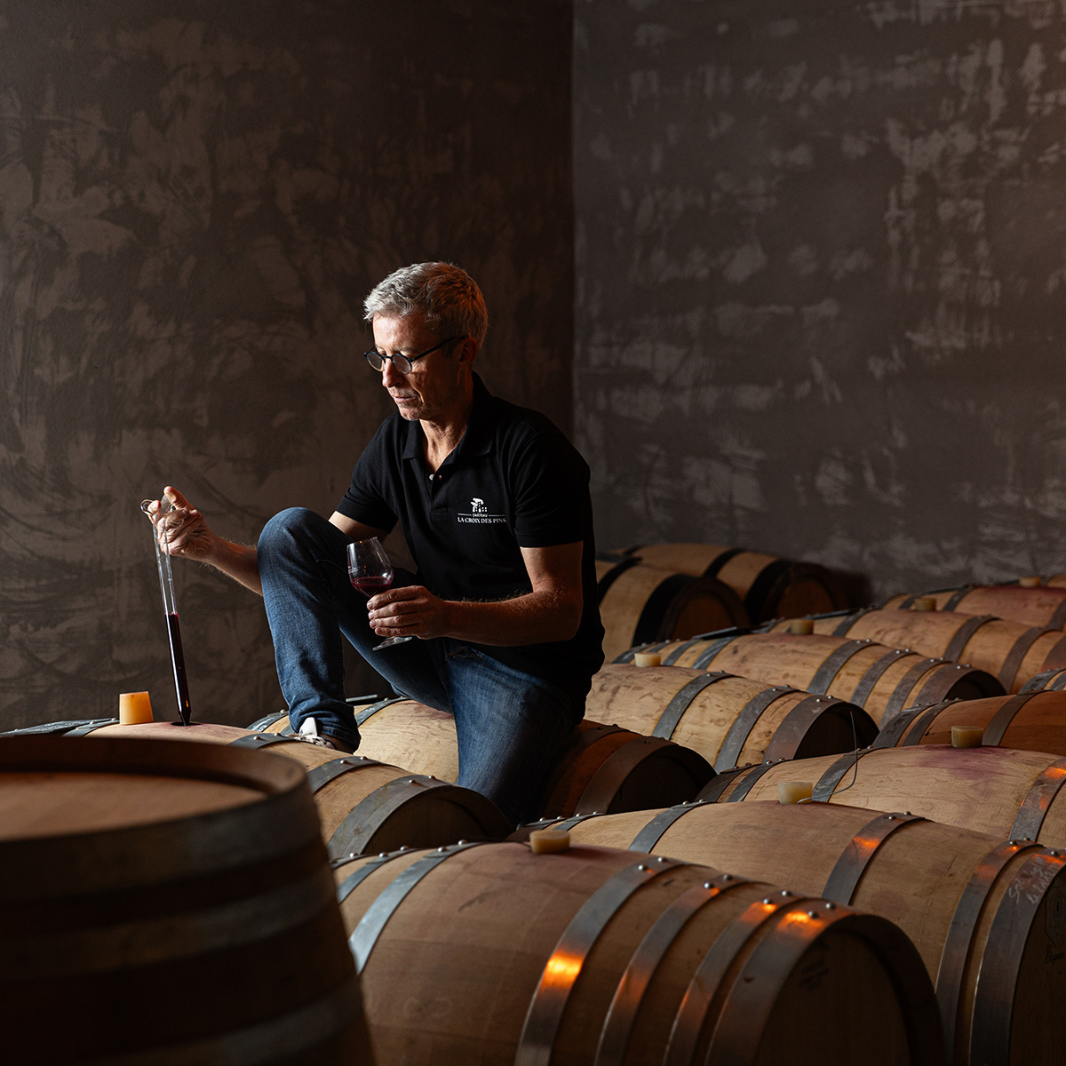 Ein Winzer sitzt in einem Weinkeller auf einem Holzfass und prüft ein Glas Rotwein. Um ihn herum sind mehrere große Holzfässer zu sehen. Das Licht ist warm und hebt die Atmosphäre eines traditionellen, französischen Weinguts hervor.