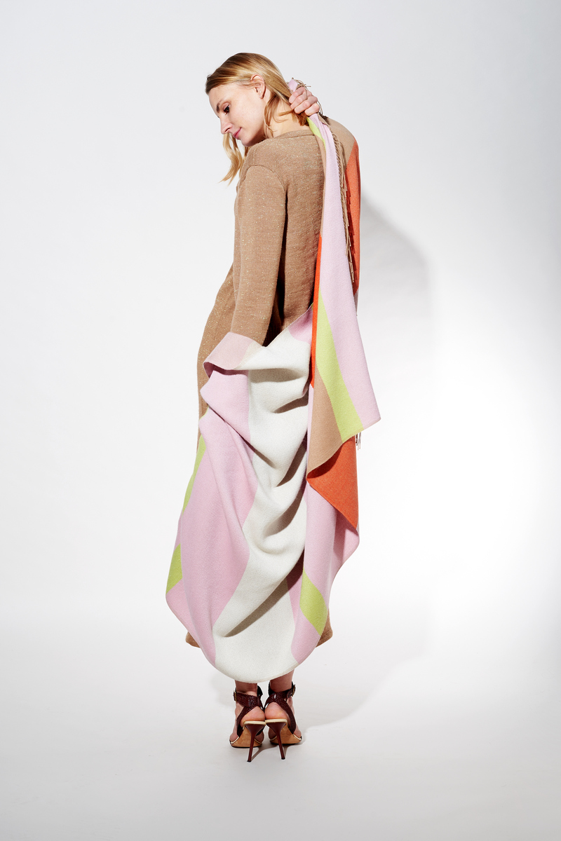 Blonde Frau trägt ein Cape aus Alpaka-Wolle in den Farben Rosa, Weiß, Grün und Orange. Die Kleidung scheint elegant und komfortabel.